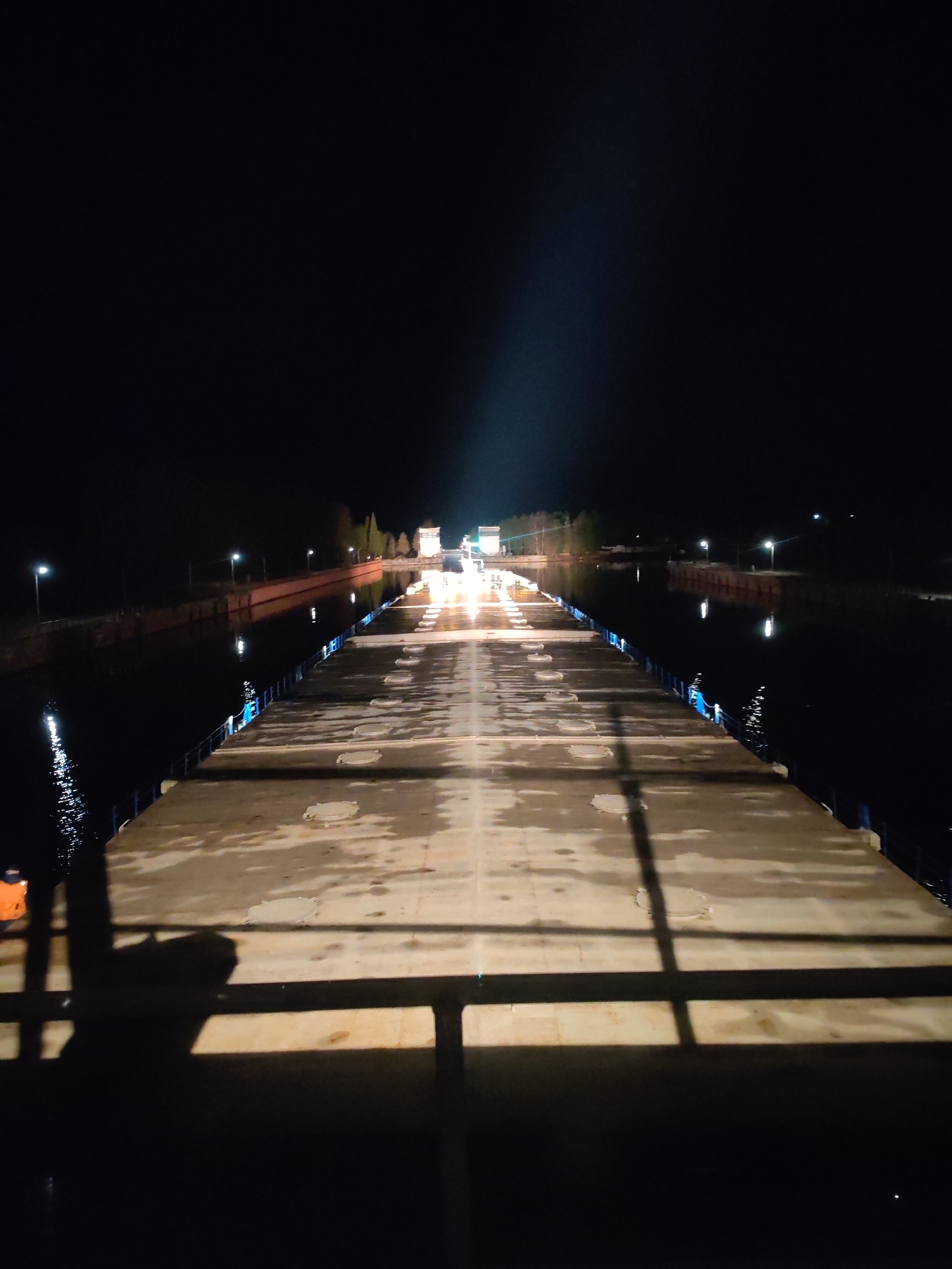A nighttime view of the Svir River Upper Lock in Svirstroy, Russia, shows a dark scene illuminated by artificial lights. The concrete surface of the lock extends towards the center of the frame, with metal railings visible along the sides and a shadowed foreground suggesting the viewer is positioned behind a barrier. The lock is damp, and illuminated by light. On the sides of the lock are still waters, reflecting the light in the dark night. Further in the distance, the lock mechanism is illuminated by a bright light, and some trees can be seen behind the lock in the background.