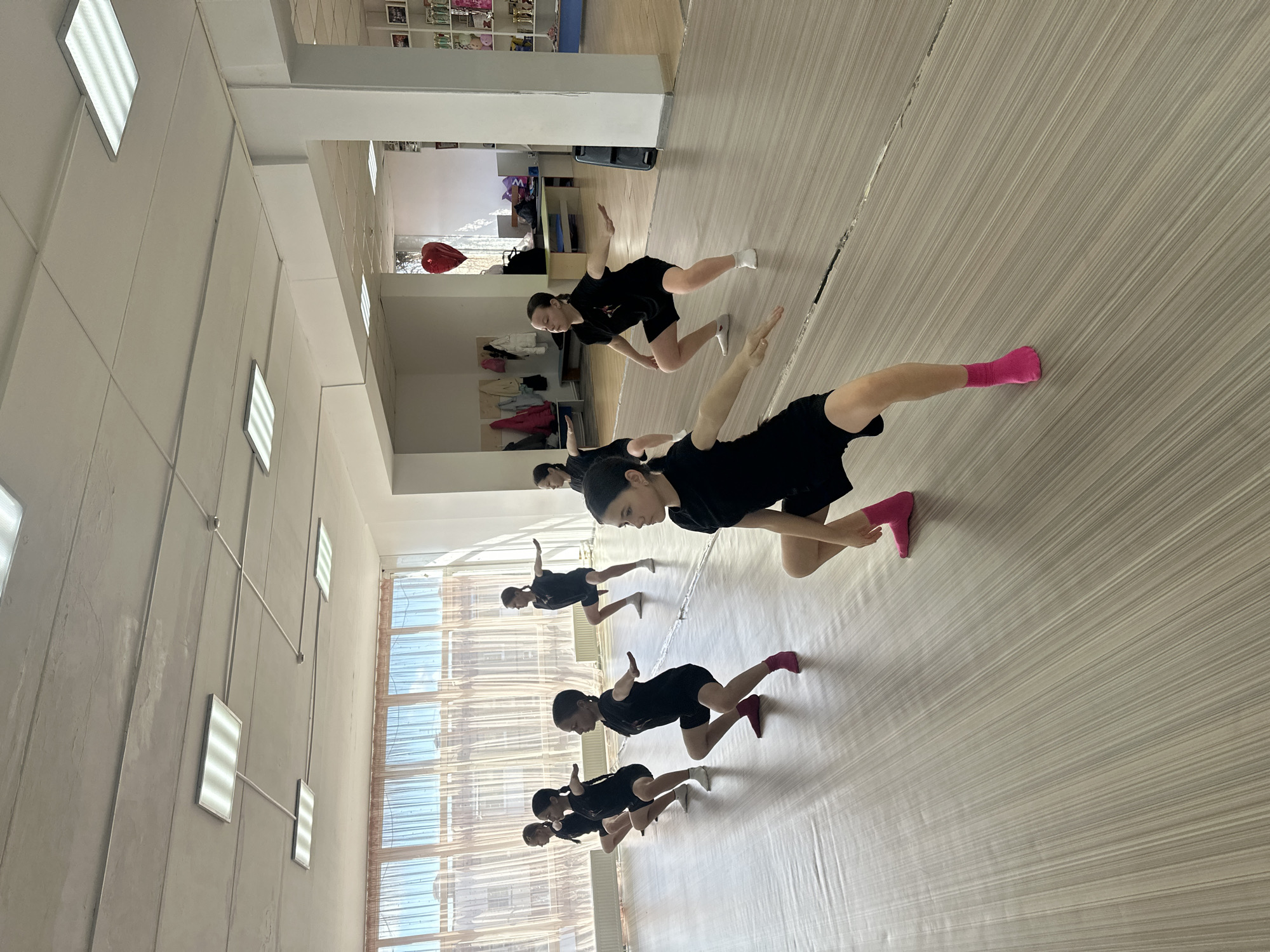 The media file captures an indoor scene featuring a group of young females, likely children or teenagers, engaged in a synchronized exercise or dance class. Six girls are visible, dressed in matching black t-shirts and black shorts. Several are wearing bright pink socks, while one has white socks. They are positioned in a line, performing a coordinated pose with their bodies bent forward, one leg extended backward, and arms outstretched for balance.
The setting appears to be a brightly lit studio or gymnasium with light-colored wooden or laminate flooring and plain light-colored walls. Large windows along one side of the room allow ample natural light to stream in, suggesting it is daytime. Rectangular fluorescent light fixtures are mounted on both the ceiling and walls. In the background, there are shelves filled with various items, possibly decorations, books, or toys. A red heart-shaped balloon is seen floating near the ceiling in the far background. The participants appear focused on their routine. There is no visible text in the image. The location is Votkinsk, Russia.