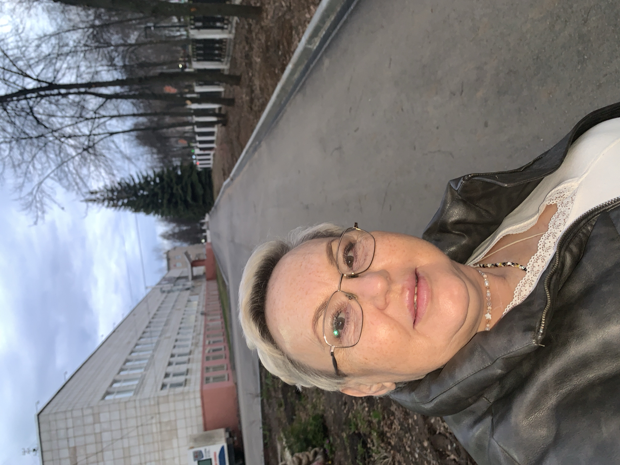 The image is a selfie of a middle-aged woman with short blonde hair and glasses, wearing a dark leather jacket over a white lace-trimmed top and a colorful beaded necklace. She is outside on a sidewalk next to a road. The backdrop features a large, multi-story building with rows of windows, typical of Soviet-era architecture. Trees with bare branches line the street, and a green pine tree is visible. Given the architectural style and location in Chaikovsky, Russia, the building is likely a residential or administrative structure common in Russian urban environments. The weather seems overcast.
