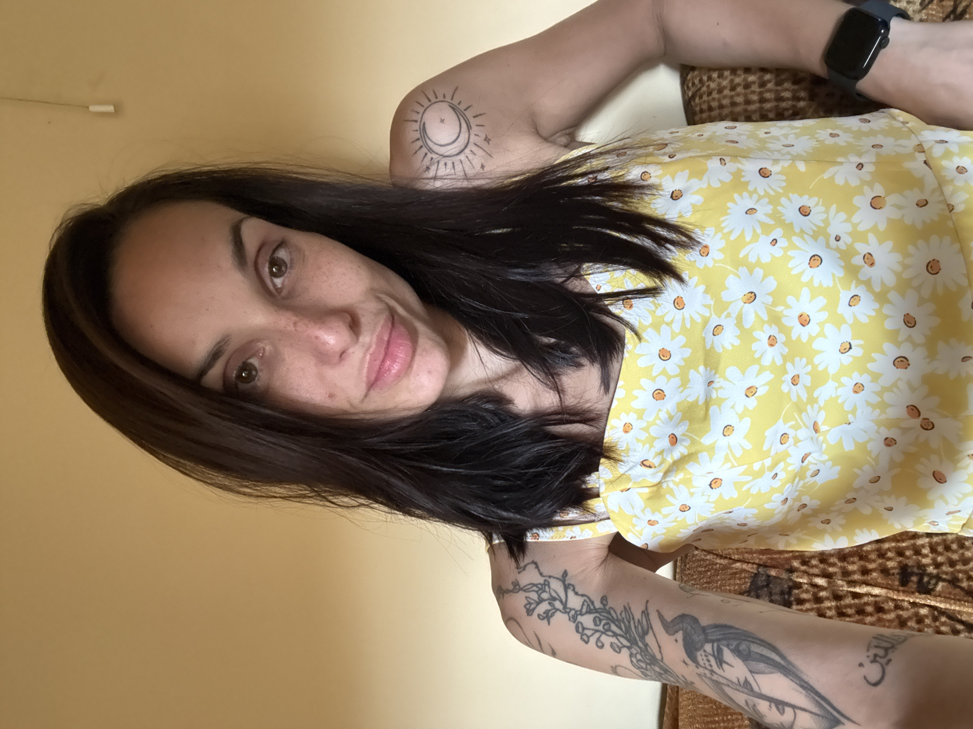 The image shows a woman with dark hair, freckles, and tattoos on her arms and shoulder. She is wearing a yellow dress with a daisy print. She is sitting on a couch with a textured brown upholstery. The background is a plain, light yellow wall with a white light switch visible. The image appears to be taken indoors, likely in a residential setting. There is no information to suggest the location is in Pleven, Bulgaria.