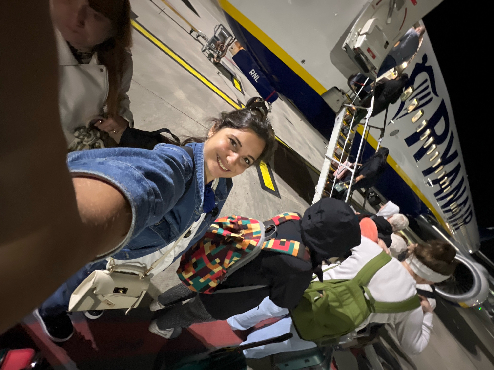 A low-angle, wide-angle shot captures a group of people boarding a Ryanair airplane on the tarmac at an airport, likely London Stansted Airport (STN) in Uttlesford, United Kingdom. The woman in the foreground, facing the camera, is taking a selfie. She has dark hair pulled back and is wearing a denim jacket. To her left, another woman with long brown hair is partially visible, wearing a white jacket. Behind the selfie-taker, several other individuals are seen ascending the airstairs of the aircraft. The airplane, with its distinctive yellow and blue livery and "RYANAIR" logo, is a prominent feature. The ground beneath is asphalt, marked with yellow lines. The scene appears to be at night or in low-light conditions, as indicated by the artificial lighting.