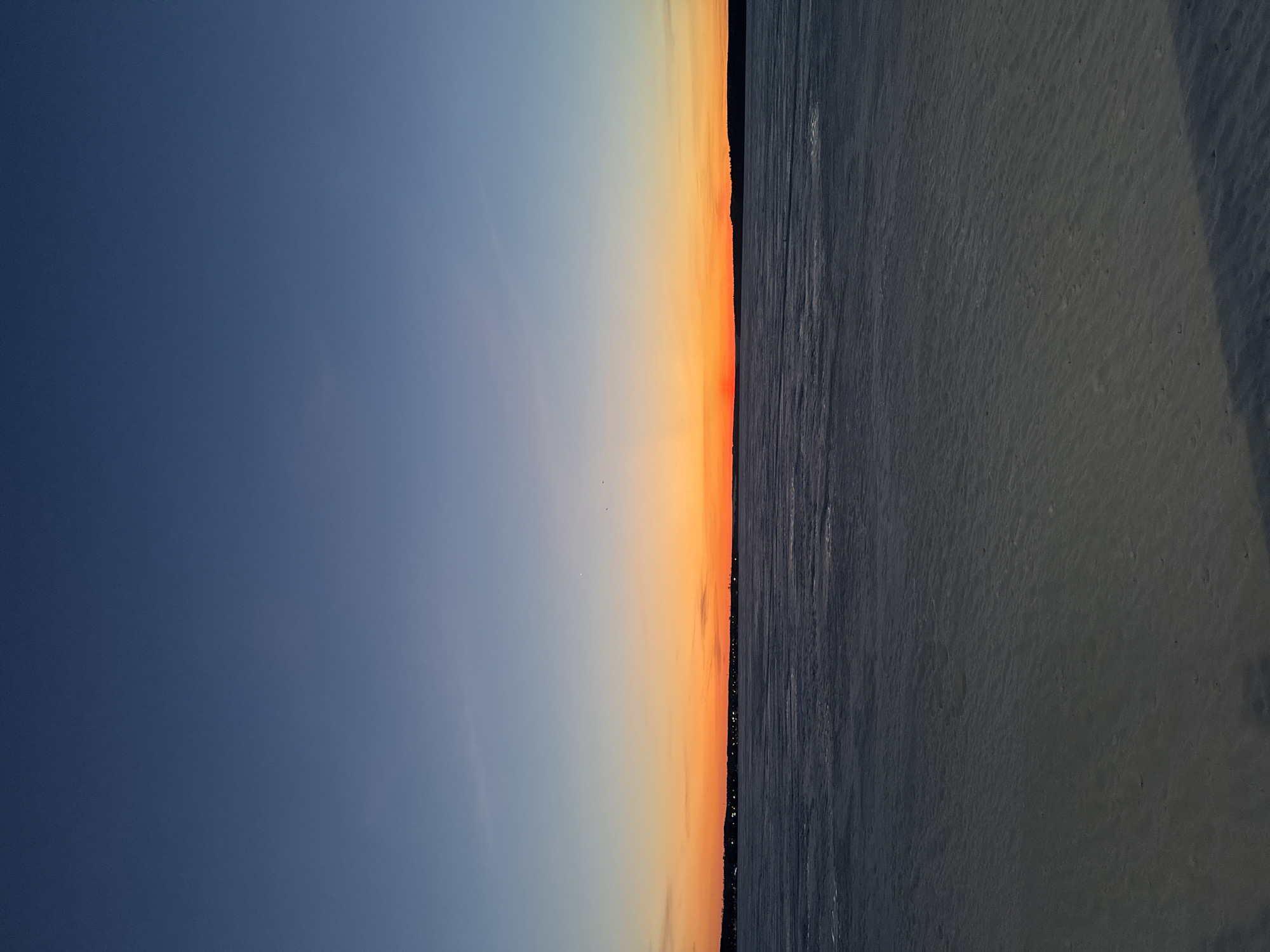 The image shows a sunset over what appears to be ice or frozen water. On the left is a dark blue sky that fades to a lighter blue, then to an orange-red sunset horizon. On the right of the horizon line is a dark gray or black surface that appears to be ice or frozen water. There is a dark line that could be land or trees on the horizon, between the sunset and the ice. The location could be Votkinsk, Russia, based on the frozen landscape possibly being a reservoir or lake in winter. There are no people in the frame.