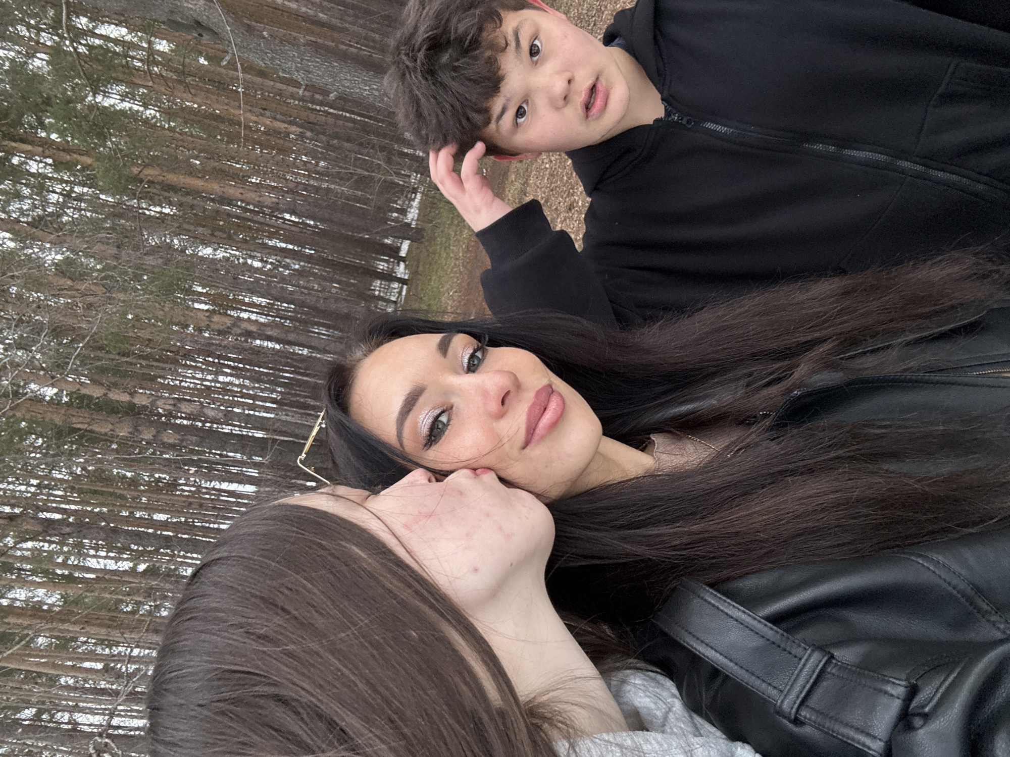 Three people are in a wooded area. A young woman with dark hair is in the center of the frame and looks at the camera. Another young woman with dark hair is to the left of her. The left young woman is angled toward the central woman, appearing to kiss her cheek. A young boy with dark hair is behind the central woman and looks at the camera with a questioning expression and a hand on his head. All three people are wearing dark clothing. The location is likely outdoors, a wooded area, as evidenced by the many bare trees visible in the background. The location is the city of Chaikovsky, Russia. The ground appears to be covered with leaves and snow.