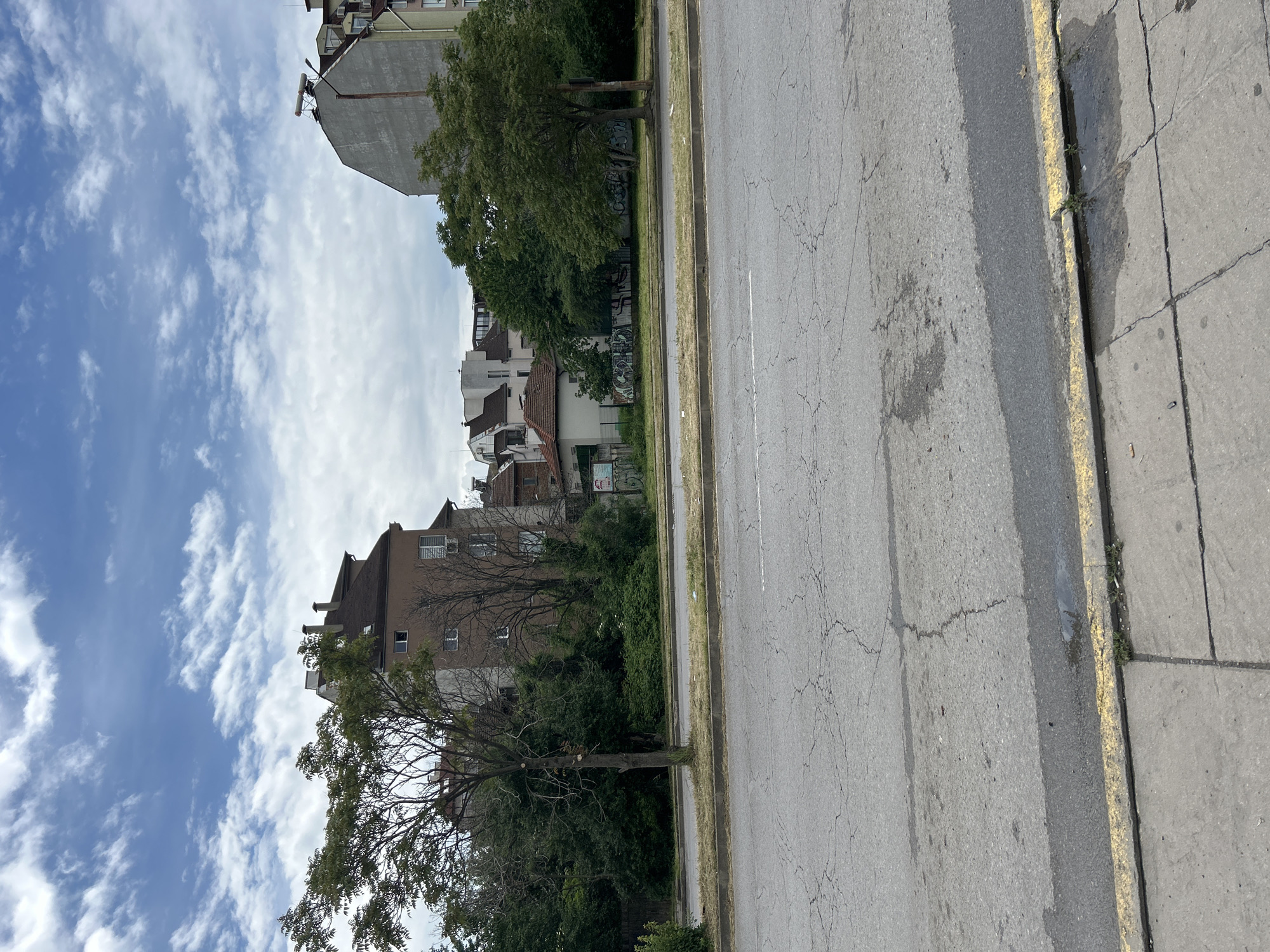 The image depicts a street scene in an urban area of Unknown, Unknown. A cracked asphalt road runs parallel to a sidewalk with a yellow-painted curb. Buildings of varying heights and architectural styles line the opposite side of the road, interspersed with trees and vegetation. The sky is blue with scattered clouds. No people are visible in the frame.