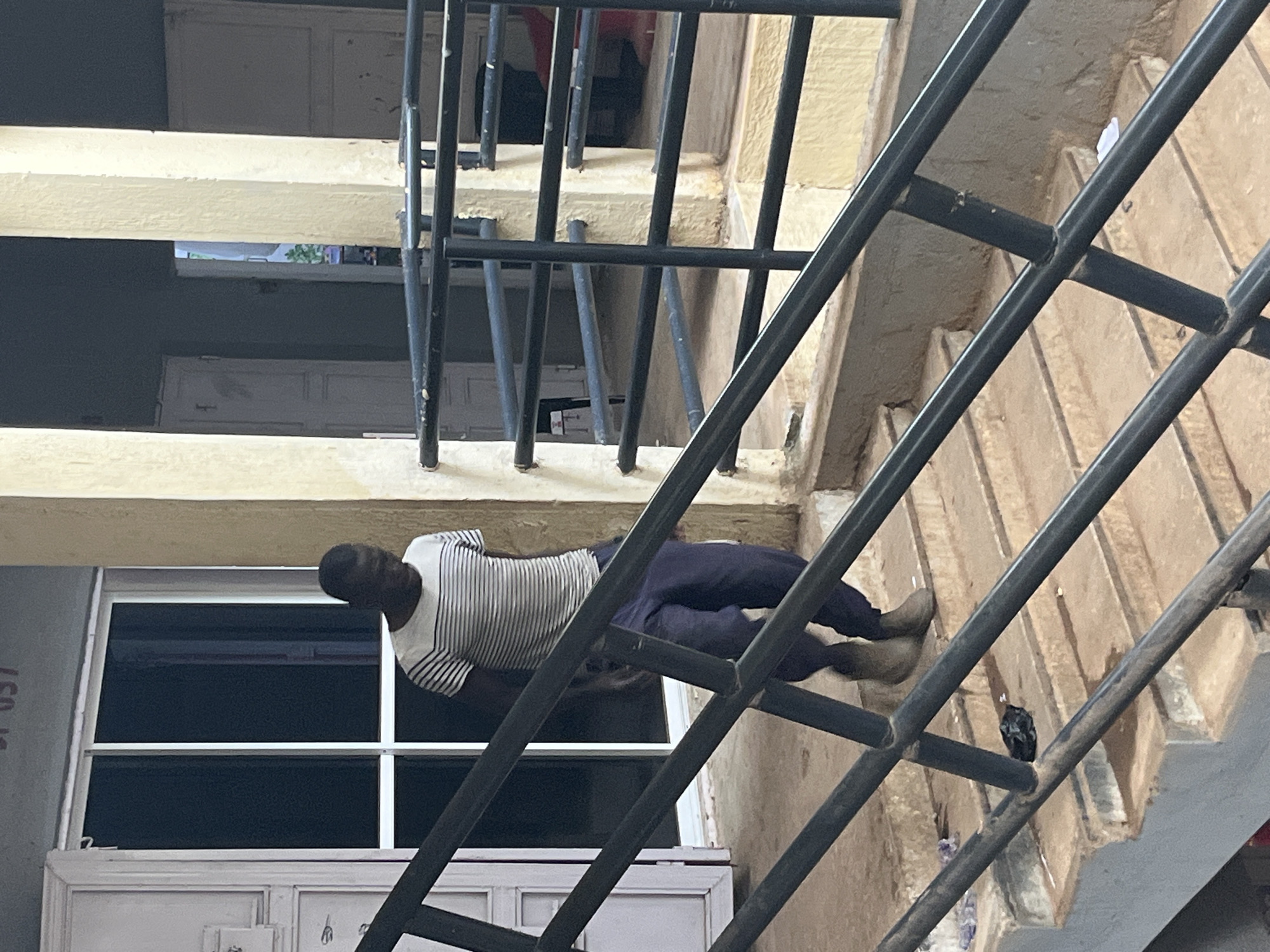 A dark-skinned man wearing a horizontally striped t-shirt and blue trousers walks down a concrete staircase with a black metal railing. He is in an outdoor setting, possibly in the city of Jos, Nigeria, characterized by weathered concrete pillars and an open entryway with a door. The staircase is partially obscured by the railing. There is some debris scattered on the stairs.