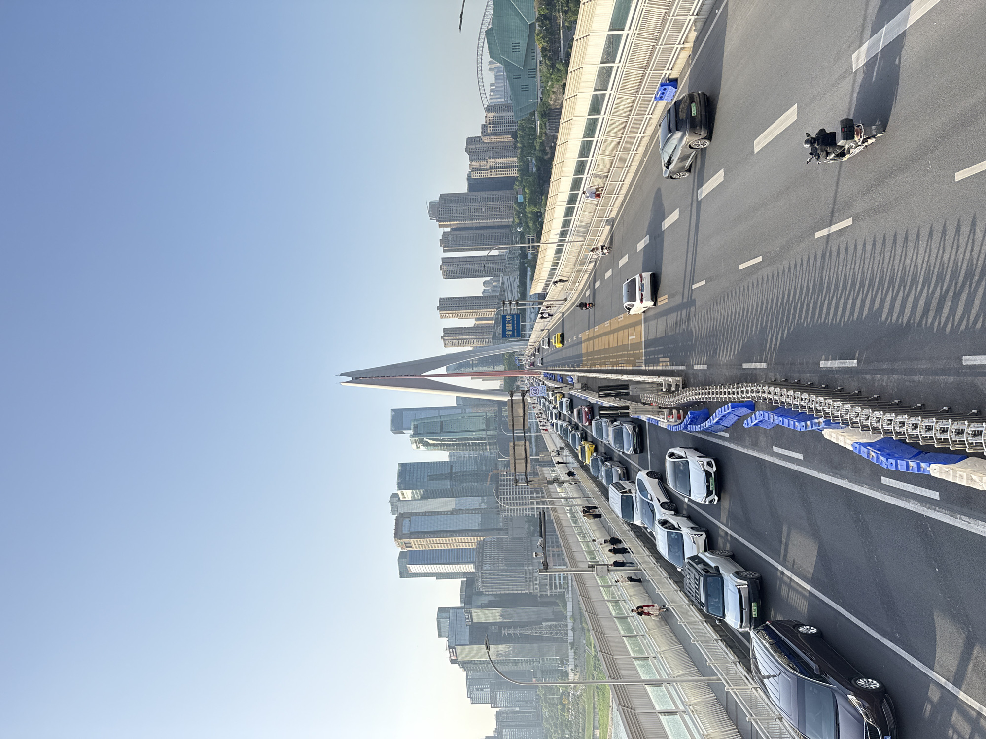 The image captures a vibrant urban scene from a bridge, looking along its length with a city skyline in the background. The perspective is from an elevated position, likely from a higher point on the bridge itself or an adjacent structure.
**Subjects:** Numerous cars of various colors (white, black, grey) are visible on the multi-lane roadway of the bridge. Some appear to be in slow-moving traffic, while others are spaced out. At least two motorcycles are also present on the road. Along a dedicated pedestrian walkway on the left side of the bridge, several people are seen walking. The background is dominated by a dense cluster of modern high-rise buildings and skyscrapers, signifying a bustling city. A prominent, white cable-stayed bridge pylon rises in the distance on the left side of the bridge structure.
**Activity or event taking place:** The scene depicts daily urban traffic and pedestrian movement on a major bridge. Vehicles are commuting, and people are walking on the pedestrian path.
**Notable details:** The time of day appears to be either morning or late afternoon, characterized by long, distinct shadows cast by vehicles and bridge structures onto the asphalt road. The sky is clear and a pale blue, indicating sunny weather. Blue and white plastic barriers line sections of the roadway, possibly for traffic management or an event. The bridge itself features multiple lanes, marked with dashed and solid white lines.
**Visible text:** A large blue road sign on the left side prominently displays "嘉华大桥" (Jiahua Bridge) in Chinese characters, with "JIAHUA BRIDGE" in English below it, and "大坪方向" (Daping direction). A circular speed limit sign indicating "60" km/h is also visible on the road. Chinese license plates can be seen on several vehicles. The overall impression is that of a modern, well-developed urban environment in Yuzhong District, China.