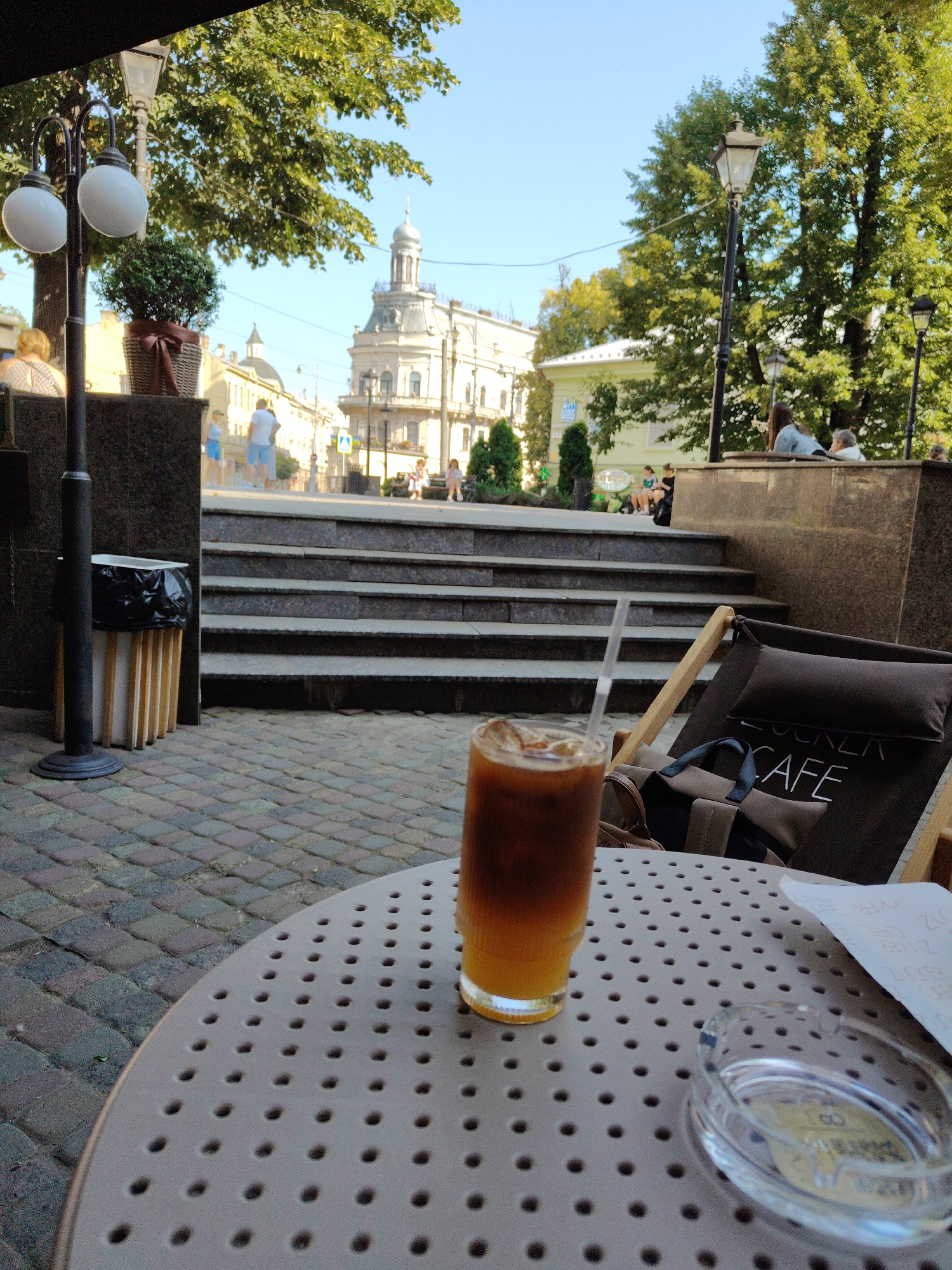 The image depicts an outdoor cafe scene with a glass of iced coffee on a table in the foreground. In the background, a street in Chernivtsi, Ukraine is visible, featuring historic architecture, trees, and people.
Specifically, the foreground shows a round, gray table with a pattern of holes. A tall glass filled with iced coffee, featuring layers of brown and orange liquid and ice cubes, stands on the table, with a clear straw inserted. Next to the glass is a glass ashtray with a coin inside. A piece of paper with some writing is also on the table. A dark canvas deck chair with a pillow is placed behind the table, bearing the word "CAFE" and some other text.
The middle ground reveals a set of stone steps leading up to a raised area where people are seated. A black trash can with a wooden base is located to the left of the steps. A lamppost with two globes is also visible on the left side of the frame.
The background shows a street lined with buildings, including a prominent building with a dome and ornate architecture, characteristic of Chernivtsi's historical center. Trees with green foliage line the street, and several people are walking or sitting in the distance. The sky is clear and blue. The overall scene suggests a sunny day in a vibrant urban setting.