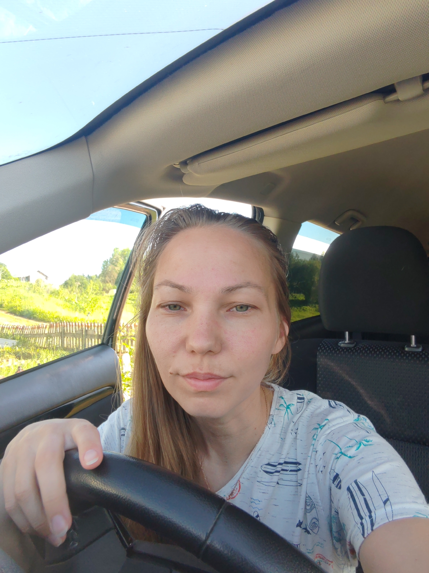 A medium shot selfie shows a woman sitting in the driver's seat of a car. The woman is facing the camera, and her left hand is on the steering wheel. She has long blonde hair, green eyes, and light skin. She is wearing a white t-shirt with a pattern of surfboards and palm trees. The interior of the car is visible, including the dashboard, steering wheel, and the back of the passenger seat. The background shows a rural landscape with green fields, trees, and a wooden fence. The sky is blue and clear, indicating a sunny day. The location is likely in a rural area, possibly outside the city of Chukhlem, Russia, given the context. The image is taken from a slightly low angle.