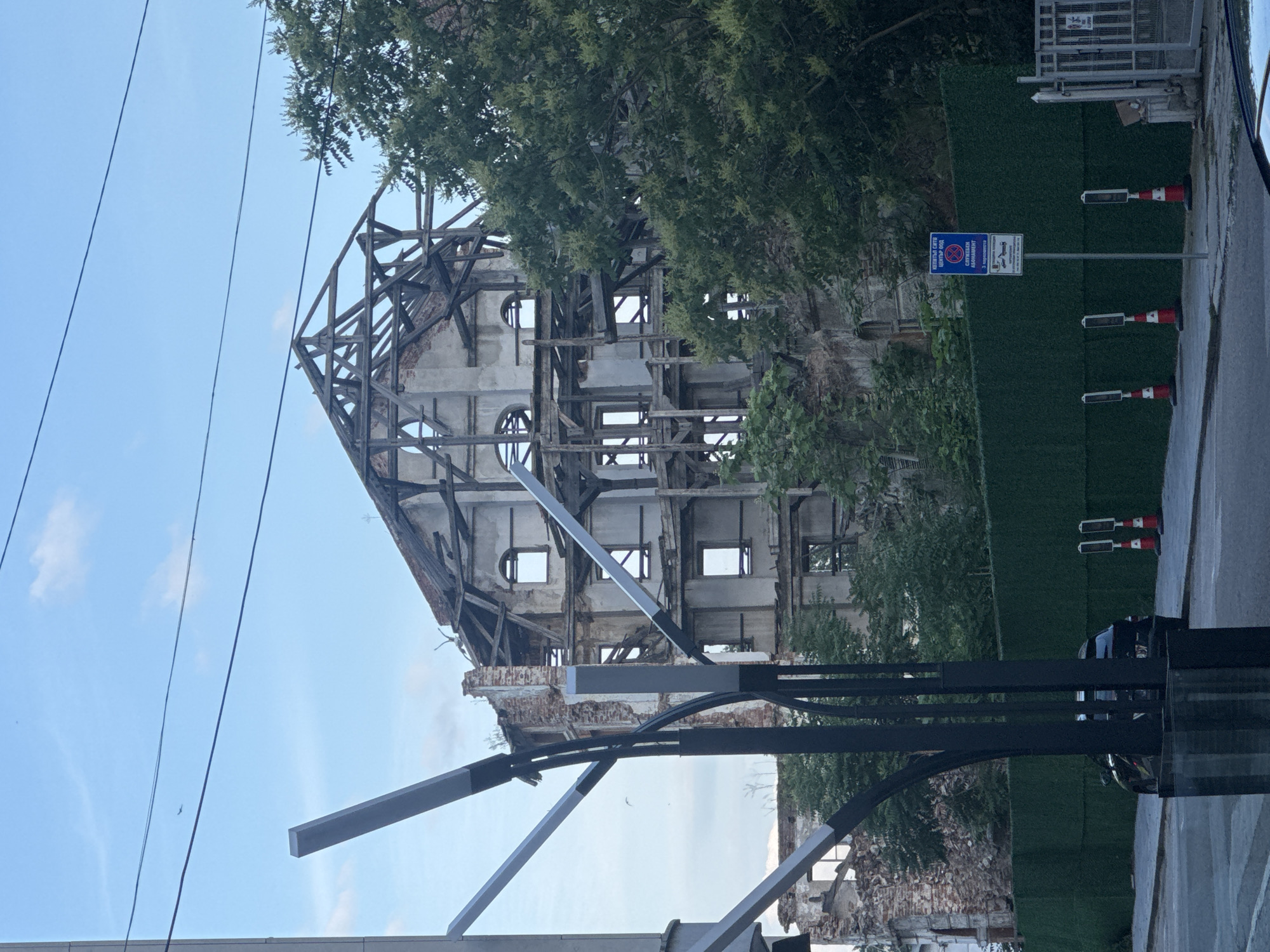 An outdoor, daytime shot captures a large, partially deconstructed or renovated building under a bright blue sky with scattered clouds. The structure appears to be an old, multi-story edifice, now largely reduced to its internal skeleton of dark wooden beams and some remaining concrete and brick walls. Several empty window frames, including distinctive round openings, are visible. Green foliage from trees partially obscures the upper left and right sides of the building, and a green mesh fence runs along the right side at ground level.
In the foreground, a modern black and grey street light structure extends diagonally across the lower half of the image. Overhead power lines traverse the sky from left to right. To the right of the building, on the side of a road, there's a blue informational sign mounted on a pole, accompanied by two red and white traffic cones. The sign, written in Cyrillic, indicates a "TEMPORARY STOP CENTER-SOUTH" and specifies that the site is undergoing "RENOVATION OF ARCHITECTURAL HISTORICAL MONUMENT," including "3 FLOORS + ATTIC SPACE." It also lists "DESIGNER: ARCH. DIMITAR ALEKSIEV." A small section of a road or paved area is visible on the far right, with what appears to be part of a parked vehicle. Several small birds are seen flying against the sky. The scene suggests an urban area in Plovdiv, Bulgaria, where an old building, likely a historical monument, is being restored or rebuilt.