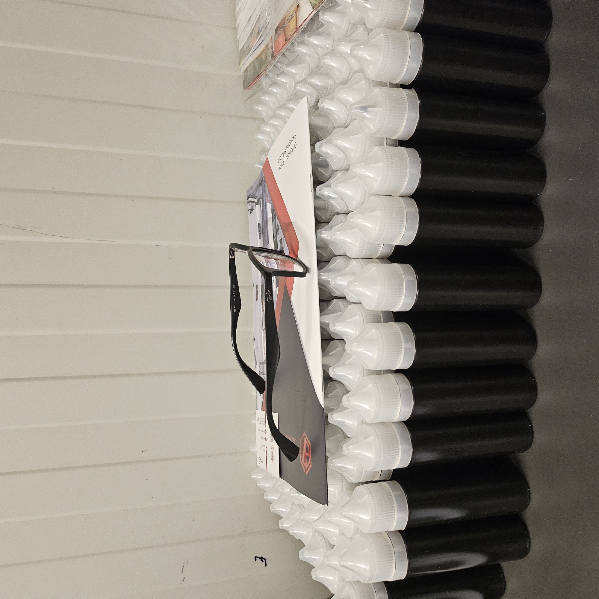 This media file captures a close-up, vertically oriented view of several rows of black plastic bottles with white caps, possibly e-liquid bottles, arranged on a dark surface. In the foreground, a pair of black-rimmed eyeglasses rests on a folded piece of paper or brochure. The brochure features red and white graphic elements and some text. Behind the bottles and to the left, a textured, light-colored wall with horizontal grooves is visible, suggesting an indoor storage or manufacturing environment. The lighting appears to be artificial, casting subtle shadows and highlighting the smooth surfaces of the bottles. A faint "E" marking is visible on the light-colored wall in the lower left quadrant. The overall scene is static and orderly, with no people or clear indications of the time of day or weather. The text on the brochure is partially legible and appears to include phrases like "Packaging Solutions" and "Customized Flexible," along with what seems to be a company logo. The location context is identified as Nowa Wieś Książęca, Poland.