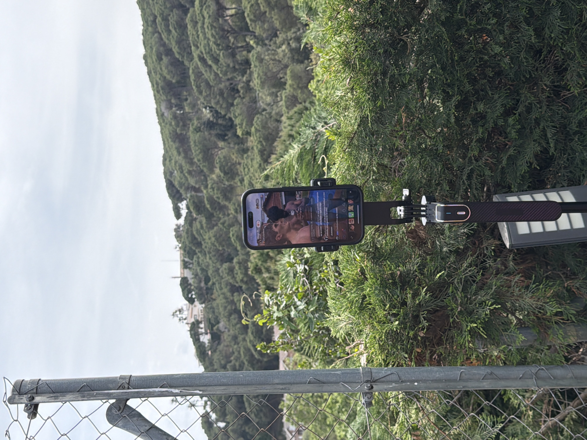 Here is a detailed description of the image:
**Scene:**
The image depicts a cellphone mounted on a device attached to what seems to be a solar lamp/light pole, positioned near a wire fence and some greenery. In the background, there is a view of buildings and trees.
**Objects:**
* Cellphone: The cellphone is mounted in a horizontal position, showing a video playing on the screen.
* Mounting Device: The device consists of an arm, a clamp holding the phone and some other connection bits.
* Solar Lamp/Light Pole: The solar lamp/light pole is positioned nearby.
* Wire Fence: A wire fence is in the foreground.
* Trees: Various trees and shrubbery occupy much of the background.
* Buildings: A few buildings are seen in the distance behind the greenery.
**Environment:**
* Location: The image appears to be taken outdoors. Based on the prompt, the location is Sant Andreu De Llavaneres, Spain.
* Lighting: The lighting is natural with some overcast conditions.
* Vegetation: Dense greenery surrounds the location.
* Weather: The sky appears overcast, suggesting a mild day.
**Summary:**
The image captures a scene where a cell phone is mounted on a pole next to a fence. The background shows the landscape with trees and buildings which is located in Sant Andreu de Llavaneres, Spain.