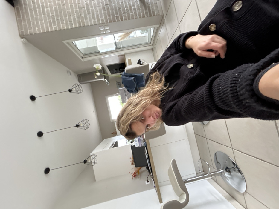 The image depicts a female subject taking a selfie inside a modern residence located in Veauchette, France. The woman, who has blonde, wavy hair, is positioned in the foreground, looking directly at the camera with her head tilted and her lips puckered. She is wearing a black, ribbed, long-sleeved garment with several large, light-colored buttons down the front. A light-colored bracelet is visible on her left wrist.
The background shows an open-plan living space featuring a kitchen area with white cabinetry and a light-colored countertop. Two contemporary bar stools with chrome bases and light-colored seats are visible. On the countertop, a dark laptop, some decorative items, a white box with the partial text "APPLELOUI", and a pair of blue jeans draped over a chair or stool are present. A white orchid plant in a dark pot is situated on an adjacent counter. A stainless steel range hood is mounted above a cooking area, and a window provides a view of greenery outside. The ceiling is white and features three black pendant lights with geometric wireframe shades. The walls are predominantly light gray, with a section of exposed light-colored brick on the right side of the frame. The floor is tiled with large, square, light-colored ceramic tiles. A large glass sliding door or window is visible on the right, reflecting indoor elements and showing some outdoor light.