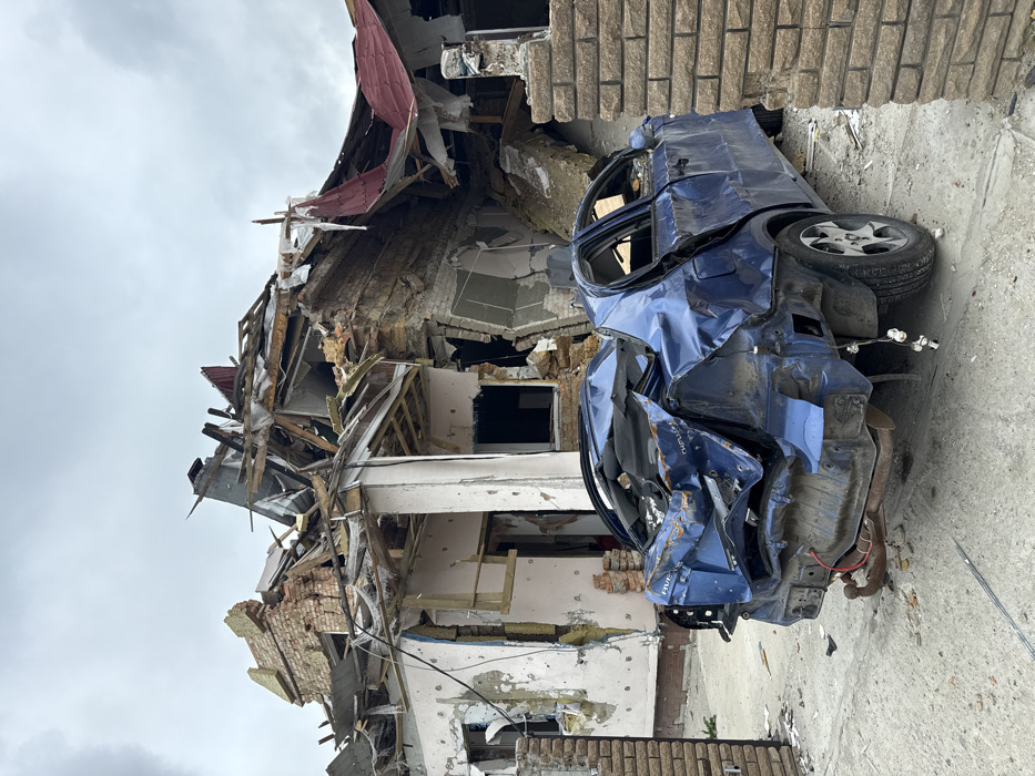 A damaged blue car sits on a cracked surface adjacent to a building in ruins. The car's body is severely crumpled, with shattered windows and missing panels. The building behind it is heavily damaged, displaying exposed brick and wooden beams. The destruction includes a collapsed roof and walls, with debris scattered around. The sky is overcast, casting a muted light over the scene.