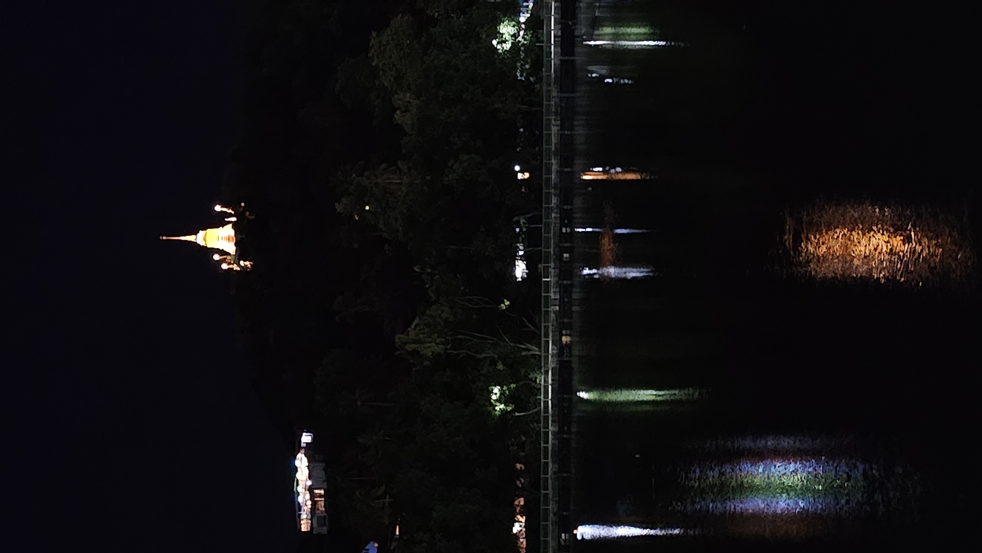 The image depicts a nighttime scene in Baan Chaweng, Thailand, characterized by artificial lighting and its reflection on a body of water. The main subject is a golden stupa, illuminated and situated on a dark, tree-covered hill. Surrounding the stupa are smaller lights, suggesting a temple complex.
To the right of the stupa, a railing or walkway is visible, also lined with lights that create distinct reflections on the water. The water itself dominates the right side of the image, serving as a mirror for the lights from the walkway and the stupa, as well as other unidentifiable lights. The reflections are distorted and elongated, creating abstract patterns of light and color on the dark surface.
The time of day is clearly night, with the darkness of the sky emphasizing the illuminated structures and their reflections. The weather appears calm, as indicated by the relatively clear reflections on the water. The scene evokes a sense of tranquility and perhaps spiritual reverence due to the prominent religious structure. There is no visible text in the image. The overall impression is one of a serene, illuminated nocturnal landscape.