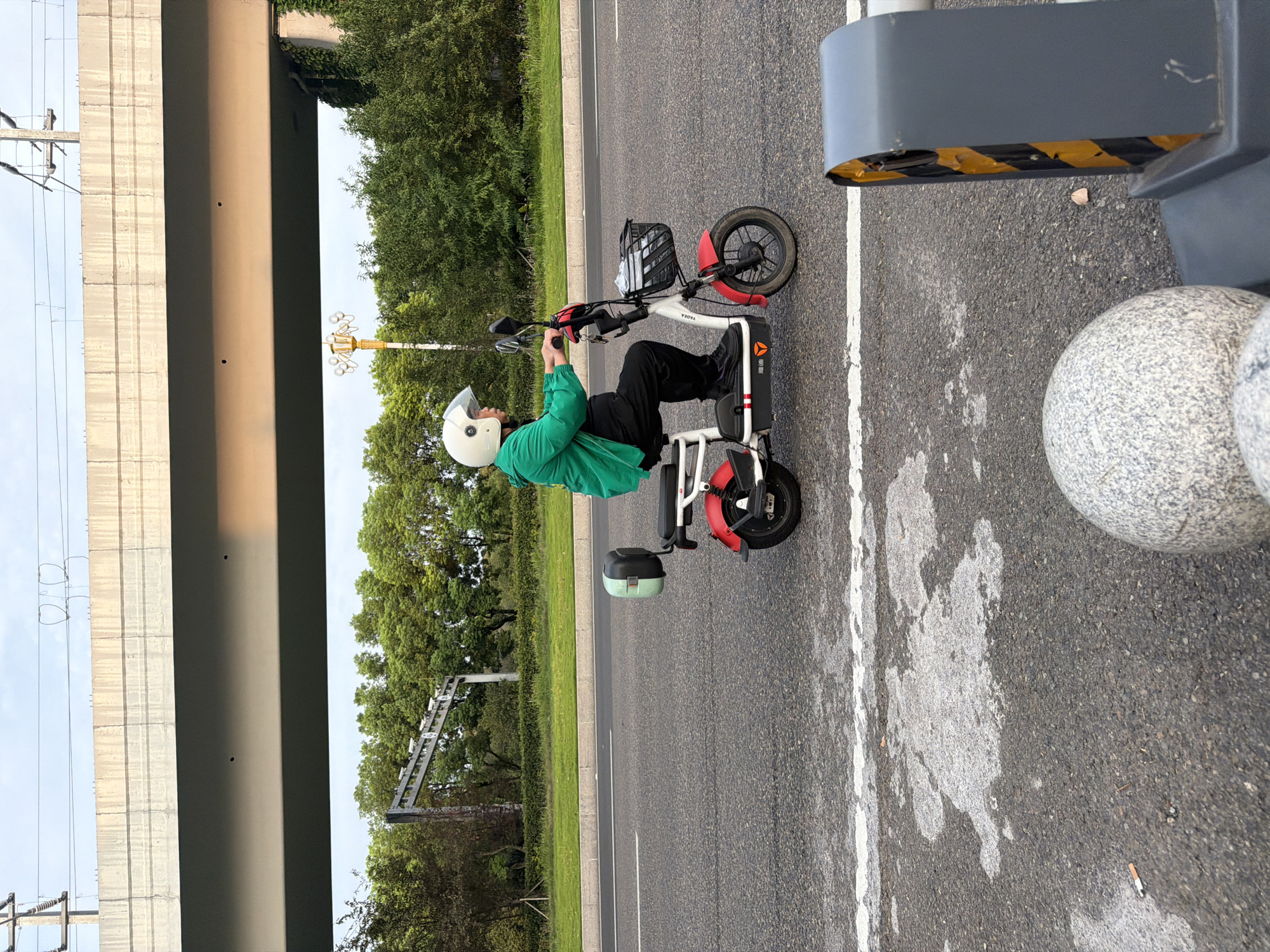 In Jiangjin, China, a person wearing a green jacket and a white helmet is riding a red and white electric scooter on an asphalt road. The scooter has a black basket on the front and a light green and black storage box on the rear. The rider is holding the handlebars and looking forward. The road has white lane markings. In the background, there are trees, a street lamp, and an elevated concrete structure, possibly a bridge or overpass, with electrical wires running overhead. The sky is overcast. In the foreground, a round granite bollard and a gray safety barrier with yellow and black stripes are visible.