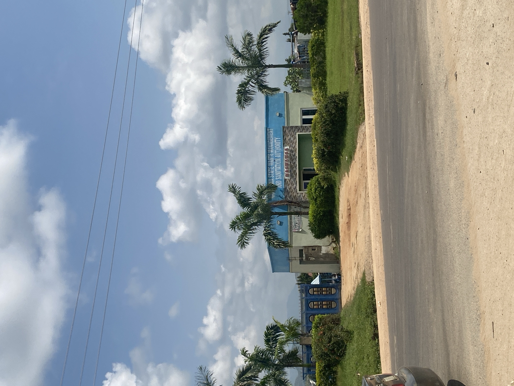 The image depicts the Niger State Waste Management and Sanitation Authority (NASWASA) building in Ado, Nigeria. The building has a light blue upper section with text visible on it and a grey lower section with green accents. A large blue gate with decorative elements is situated to the left of the building. Palm trees are scattered around the property, and the area is well-maintained with green grass and bushes. In the foreground, a road is visible with the rear of a dark-colored car parked on the left side of the frame. The sky is blue with scattered white clouds, and distant mountains are visible in the background. The scene appears to be captured during daylight.