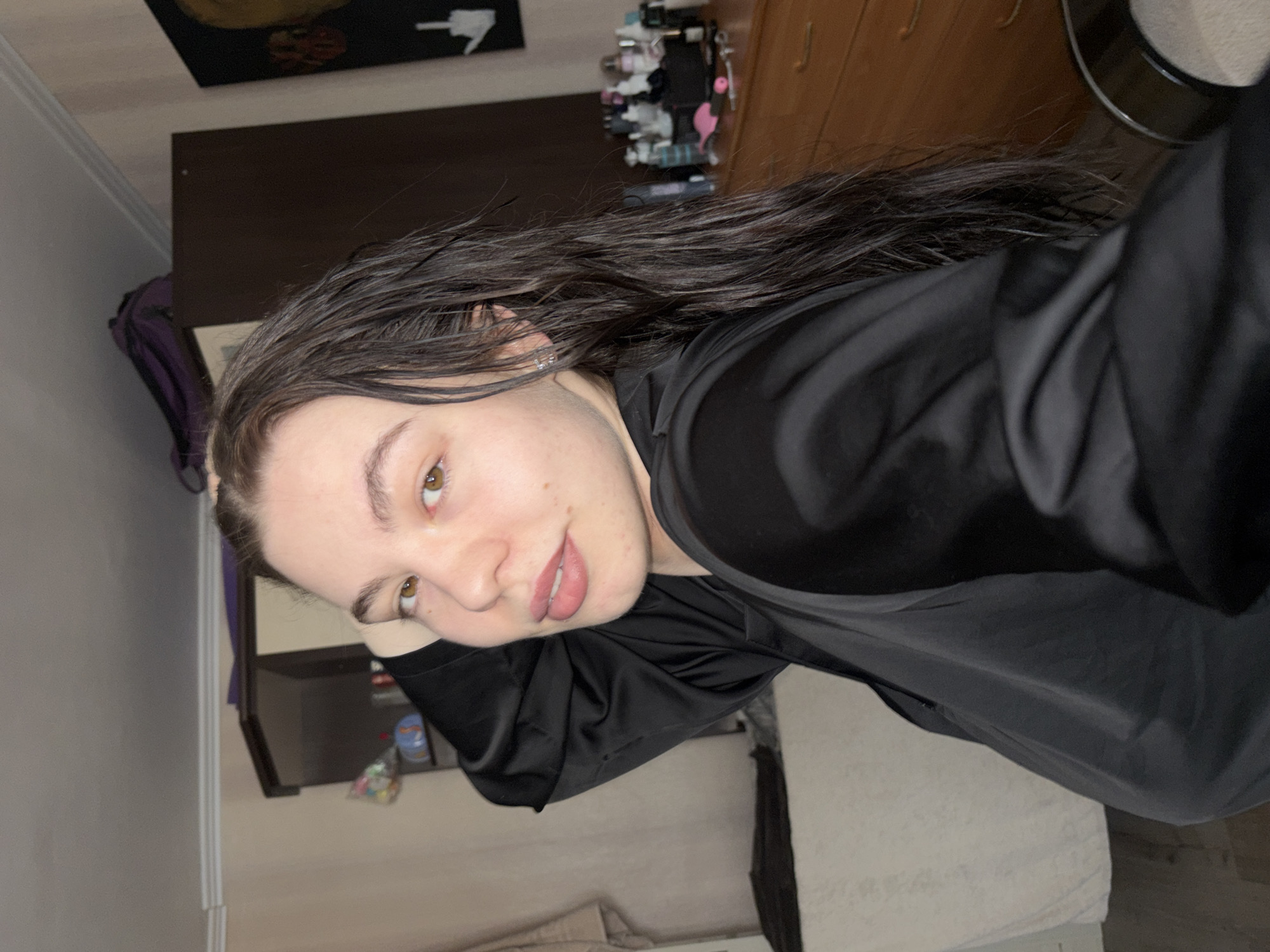 A woman with shoulder-length dark brown hair poses indoors. Her face is turned towards the viewer, revealing light skin and brown eyes. She is wearing a black satin shirt. The background shows a cluttered bedroom with a dark brown desk, various objects on top of it, and a poster on the wall. A bed with white sheets is visible in the lower part of the frame. The location is likely a residential setting but not specifically identifiable as Perm, Russia.