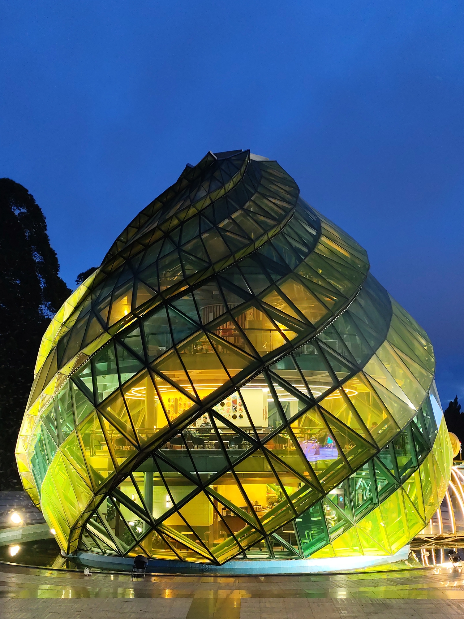 The image shows a unique architectural structure located in Da Lat, Vietnam. The building is shaped like a pine cone or artichoke, constructed with many triangular glass panels that create a spiraling effect. The glass panels are tinted in shades of green and yellow. The building is brightly lit from within, showcasing interior details such as multiple levels, lighting fixtures, and people sitting at tables. The structure sits on a slightly reflective surface, possibly water or wet pavement. The background features dark foliage, indicating the presence of trees or bushes, and a dark blue sky. The image appears to be taken at dusk or night, as the lighting is artificial and the sky is dark.