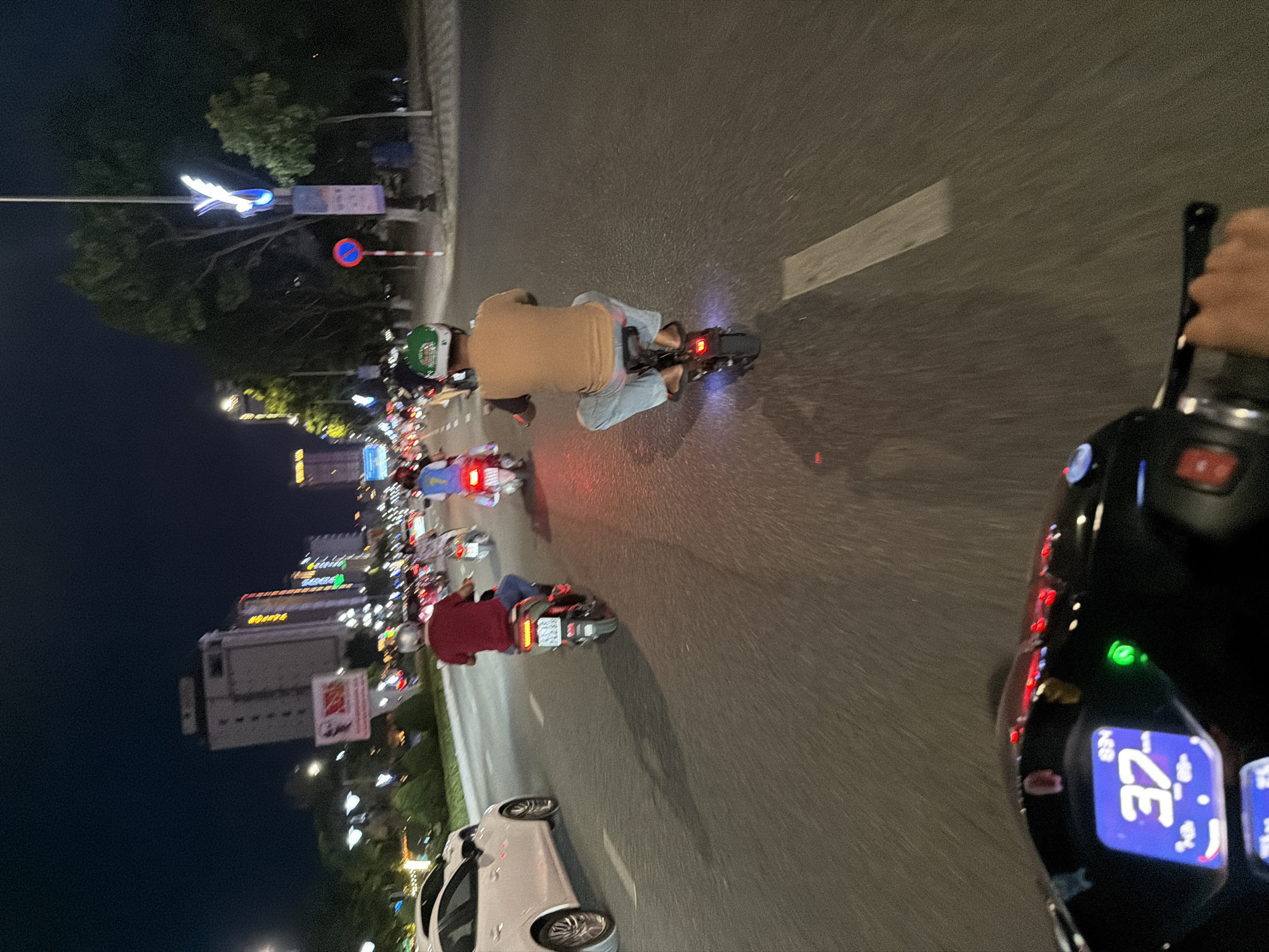 The image depicts a nighttime scene on a road in Đà Nẵng, Vietnam. The perspective is from a person riding a motor scooter, with the scooter's dashboard visible displaying a speed of "37 km/h." Ahead on the road are other scooters and vehicles. The road is illuminated by streetlights. Buildings are visible in the background, and an airplane monument on a pole. A white car is on the side of the road. The traffic appears to be moderate.