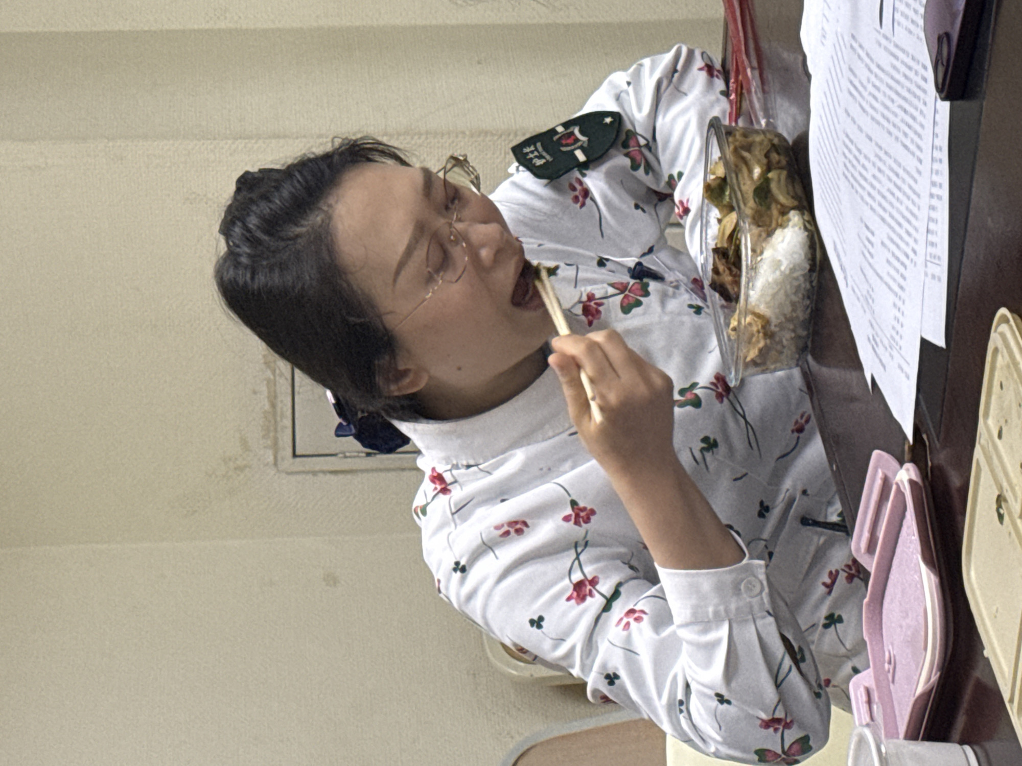 A woman with black hair and glasses is eating from a clear container with chopsticks. She is wearing a white shirt with a floral pattern and a green patch on her shoulder. The container appears to hold rice and vegetables. Papers are stacked on a table near the container. The background includes a beige wall and a window. No evidence within the image suggests a specific location in Yuzhong District, China.