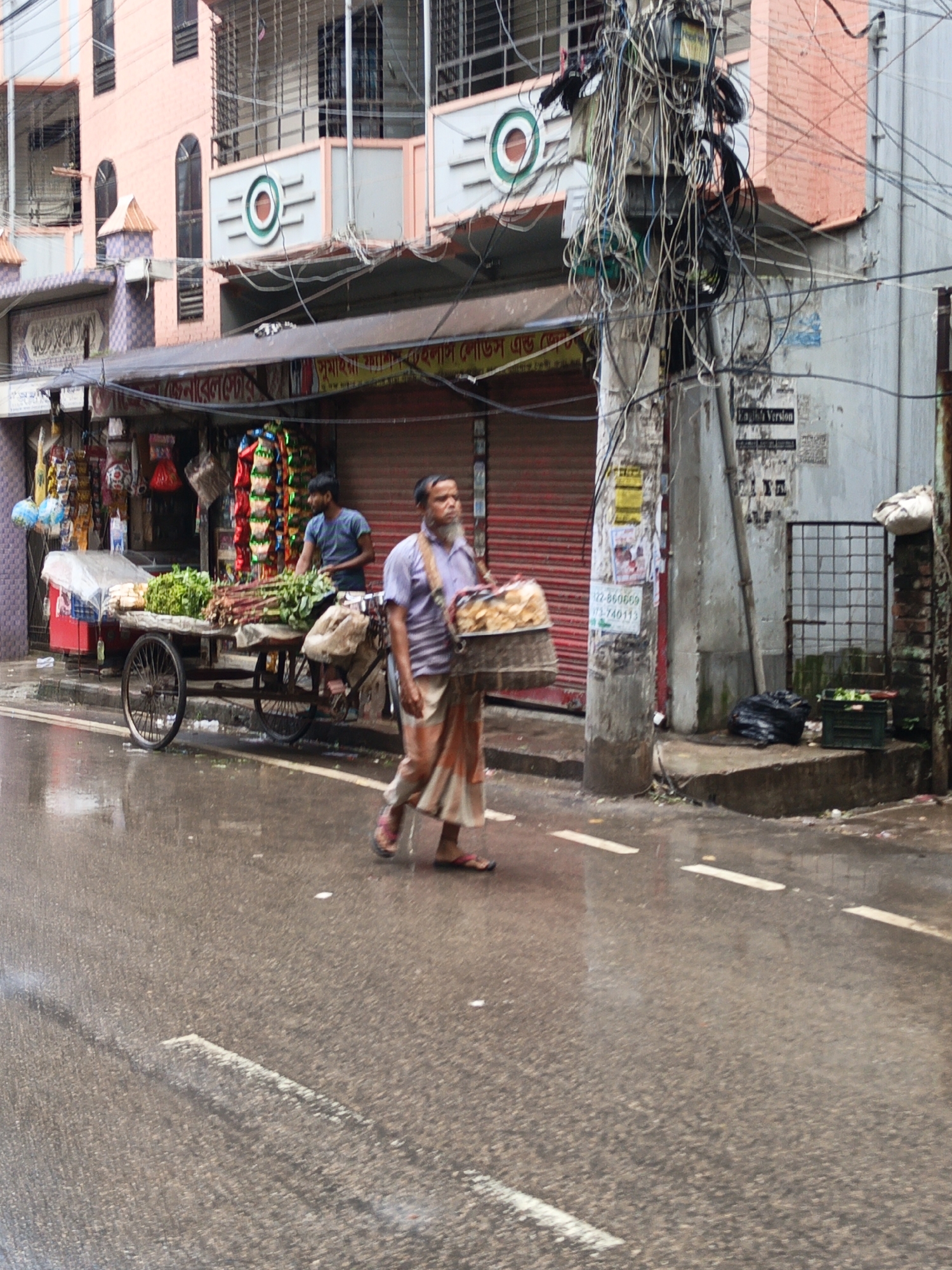 This is a medium-shot, eye-level outdoor photograph taken on a wet street in Dhaka, Bangladesh, during what appears to be daylight. The scene depicts a bustling street with a focus on a vegetable cart and two men.
In the foreground, a man with a light-colored beard and wearing a purple shirt and patterned skirt walks away from the camera, carrying a basket of goods. His feet, clad in pink sandals, are visible on the wet asphalt. The street surface is reflective, indicating recent rain.
Behind him, a vegetable cart is laden with various produce, including leafy greens and what appear to be bundles of herbs or roots. A man wearing a blue t-shirt stands behind the cart, seemingly attending to his goods. The cart has two large wheels and is positioned on the side of the street.
The background reveals a multi-story building with pink and gray facades, adorned with decorative elements. There are various shops with shuttered entrances and awnings. A prominent feature is a utility pole heavily draped with a dense network of tangled electrical wires, a common sight in Dhaka. A faded sign with Bengali script is visible on one of the shops. The overall environment suggests a commercial area within the city.
