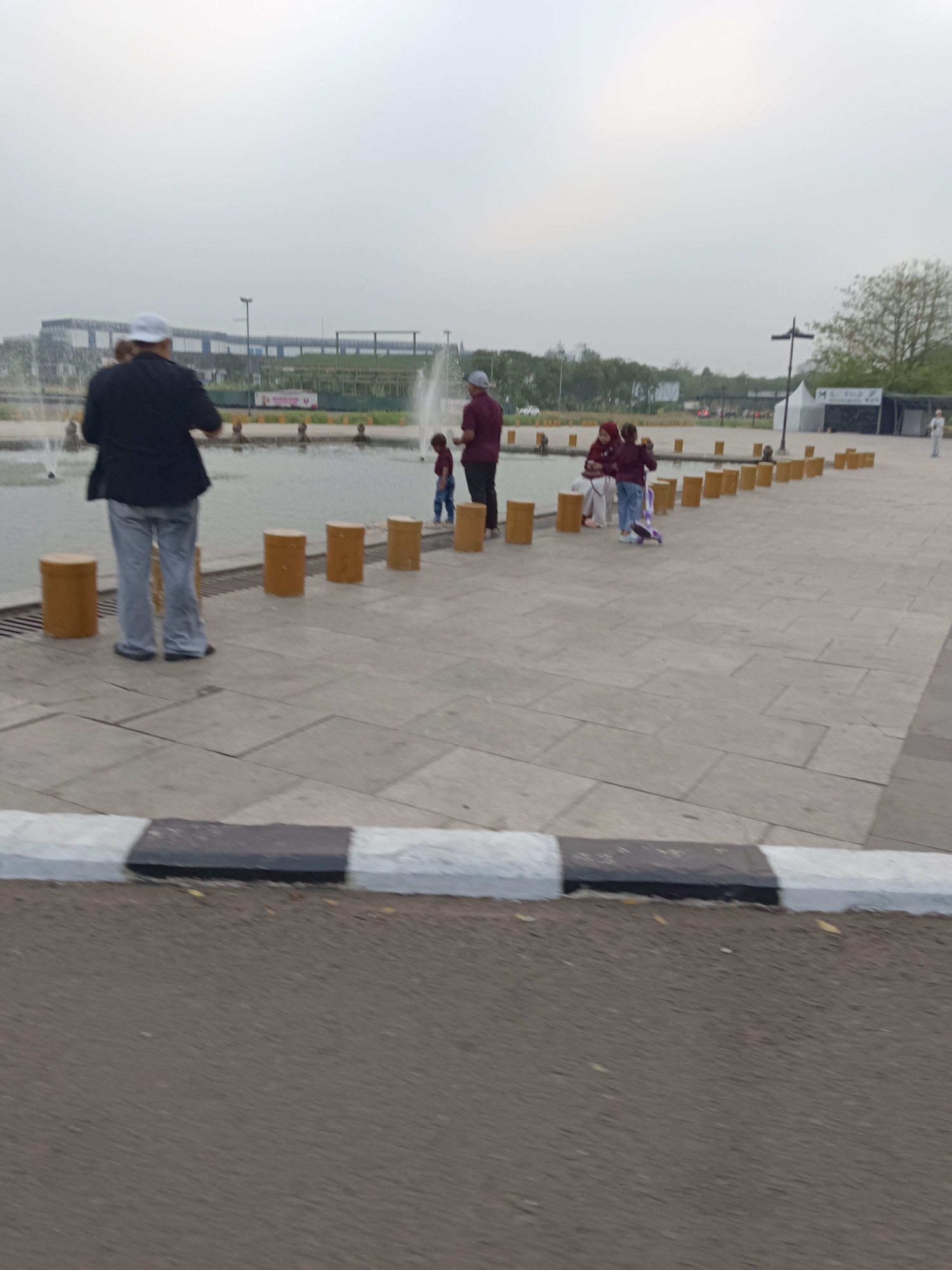 In Jayamukti, Indonesia, a paved area borders a body of water with fountains. Several cylindrical yellow posts are arranged in a line at the edge of the paved area, leading towards the water.

In the foreground, a person wearing a black jacket and light-colored pants stands facing away from the camera, looking towards the fountains. To the right of this person, a group of three people, two children and an adult, are gathered. The adult is wearing a maroon shirt and a cap, and appears to be interacting with the children. One child, in a maroon top and blue pants, is standing near the adult. Another child, also in maroon clothing, is on a scooter to the right of the first child.

Further in the background, more fountains are visible, and beyond that, a large building with a modern architectural design. The sky is overcast, suggesting a cloudy day. The paved area is made of large, rectangular tiles. A curb with alternating black and white stripes separates the paved area from an asphalt road in the immediate foreground.