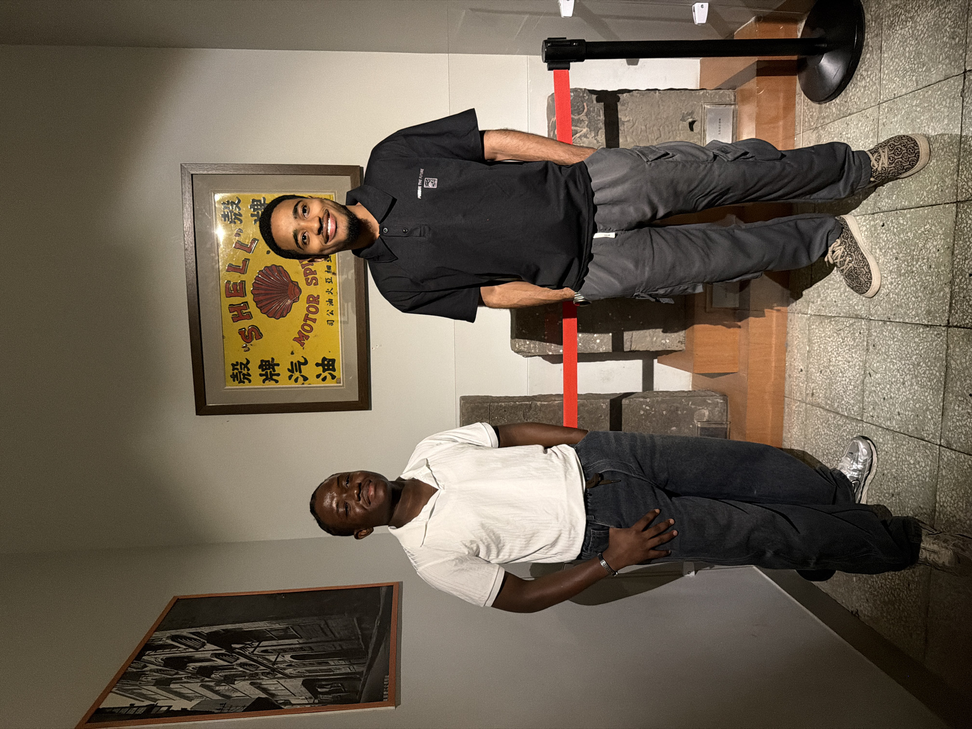 Two young men are standing in front of an exhibit within a museum located in Yuzhong District, China. The man on the left is wearing a white polo shirt and dark wash jeans, with his hands in his pockets. The man on the right is wearing a black polo shirt with a small emblem and text that reads "PURSUIT THE FUTURE", along with grey cargo pants. He also has his hands in his pockets. Behind them, a yellow framed vintage advertisement for Shell Motor Spirit is displayed, featuring Chinese characters and a red shell logo. To the left of the advertisement is a framed black and white photograph of a street scene. In the foreground, several stone structures are partially visible behind a red velvet rope barrier. The floor is tiled with a speckled pattern.