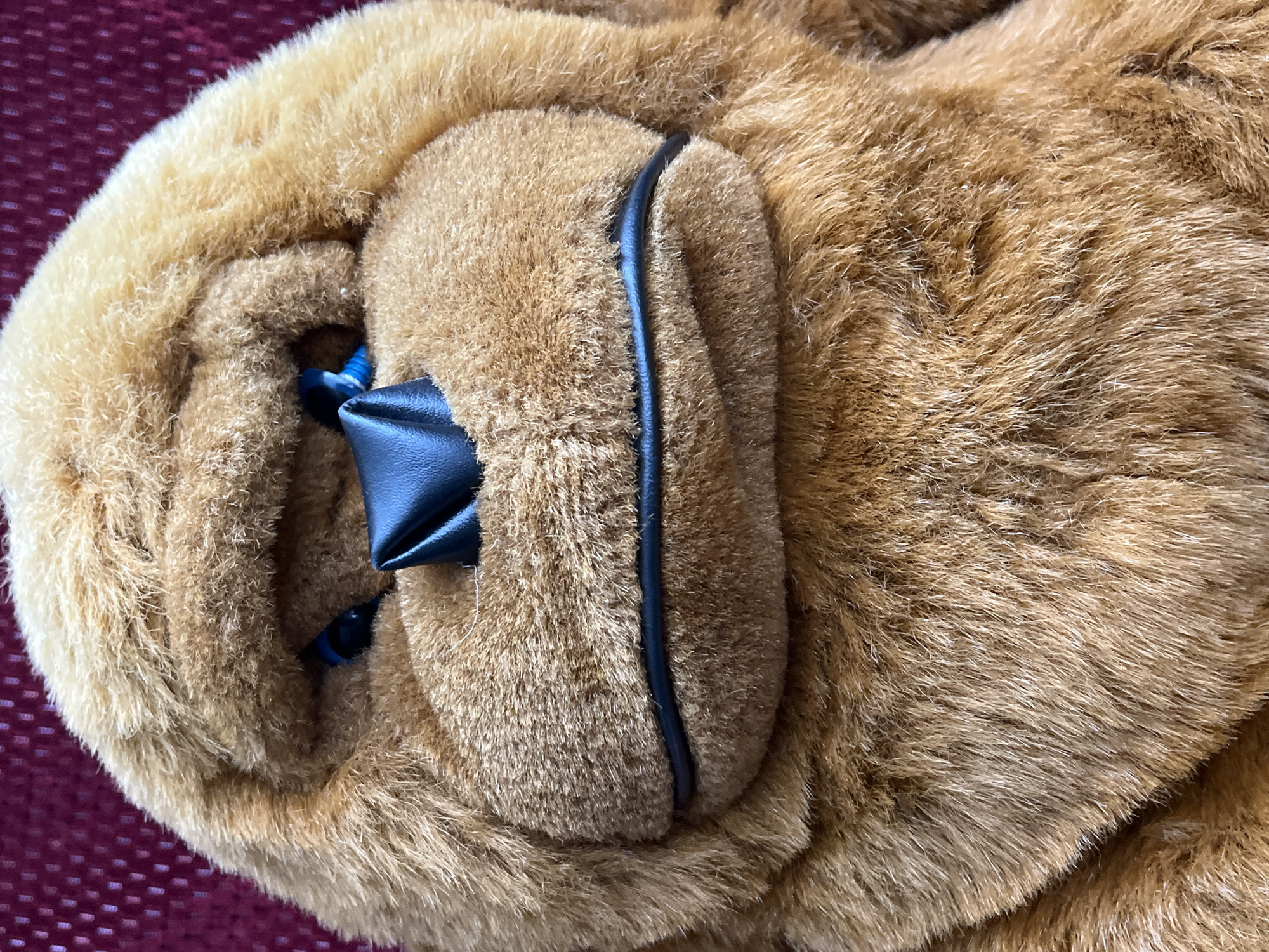 A close-up shows a light brown, furry object, possibly a stuffed animal or toy. A black, leather-like material is visible near the edge, possibly part of a decorative element. Blue accents can be seen within the fur. The background is a dark red, textured surface. The location is unknown.