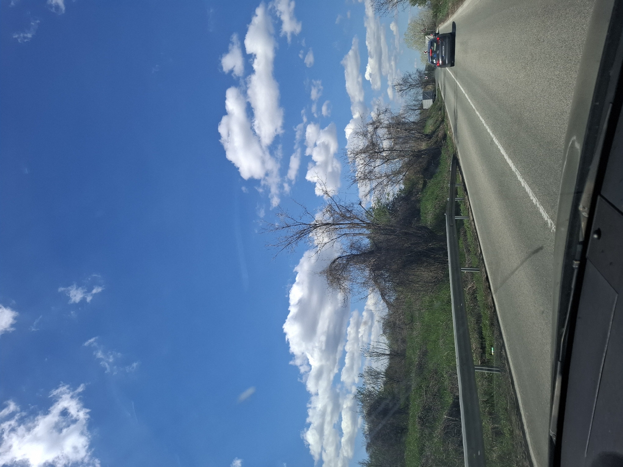 A view from inside a car, looking out onto a road in Tsar Simeonovo, Bulgaria. A dark-colored car is ahead, traveling on the asphalt road. To the left of the road, there is a grassy area with some trees. Above the scene, there is a blue sky with puffy white clouds. A metallic guardrail runs along the roadside.
