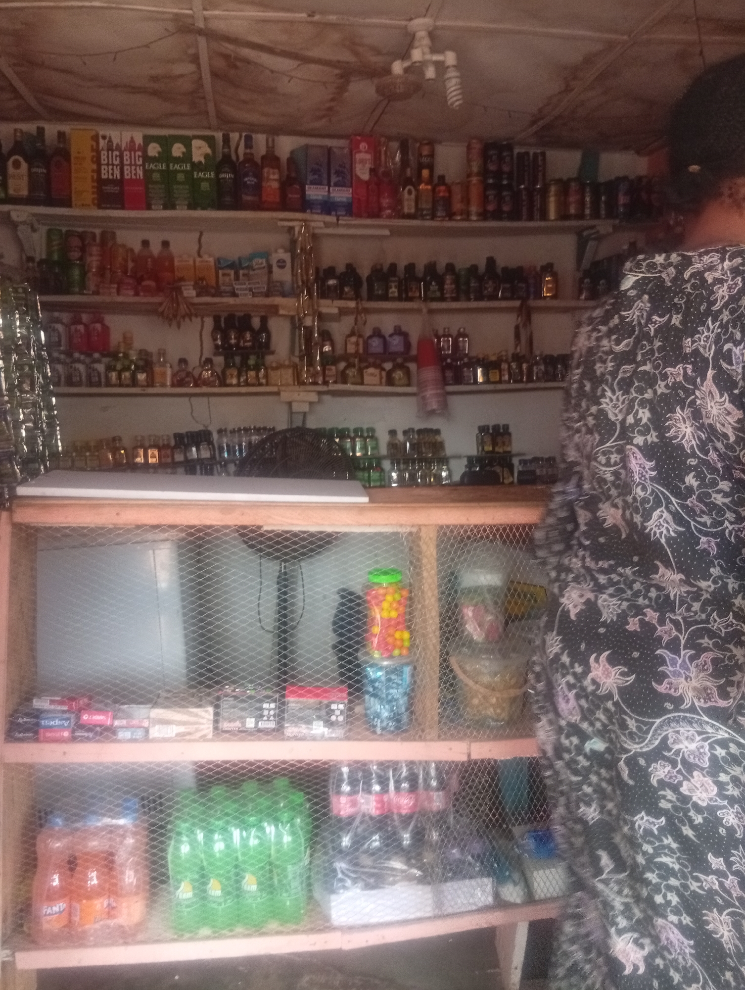 The image depicts a small, indoor retail stall, possibly a kiosk or a section of a larger market, in Yelwa, Nigeria. The stall is densely stocked with various goods displayed on wooden shelves. Prominently featured are numerous bottles of alcoholic beverages, including brands like "Big Ben," and "Eagle." Several rows of smaller bottles, likely spirits or liqueurs, are also visible. In addition to alcoholic drinks, the shelves also hold cigarettes, packaged goods, and what appear to be smaller consumer products like toiletries or condiments. On the lower shelves, there are cases of soft drinks, including Fanta and other green-bottled beverages, as well as some packaged snacks or other small items. A person, partially visible on the right side of the frame, is wearing a dark, patterned fabric garment and appears to be standing behind the counter or the display area. The stall's structure is simple, with wooden shelves and a wire mesh enclosure in the lower section. The lighting appears to be artificial, and the overall environment suggests a functional, perhaps modest, commercial space.