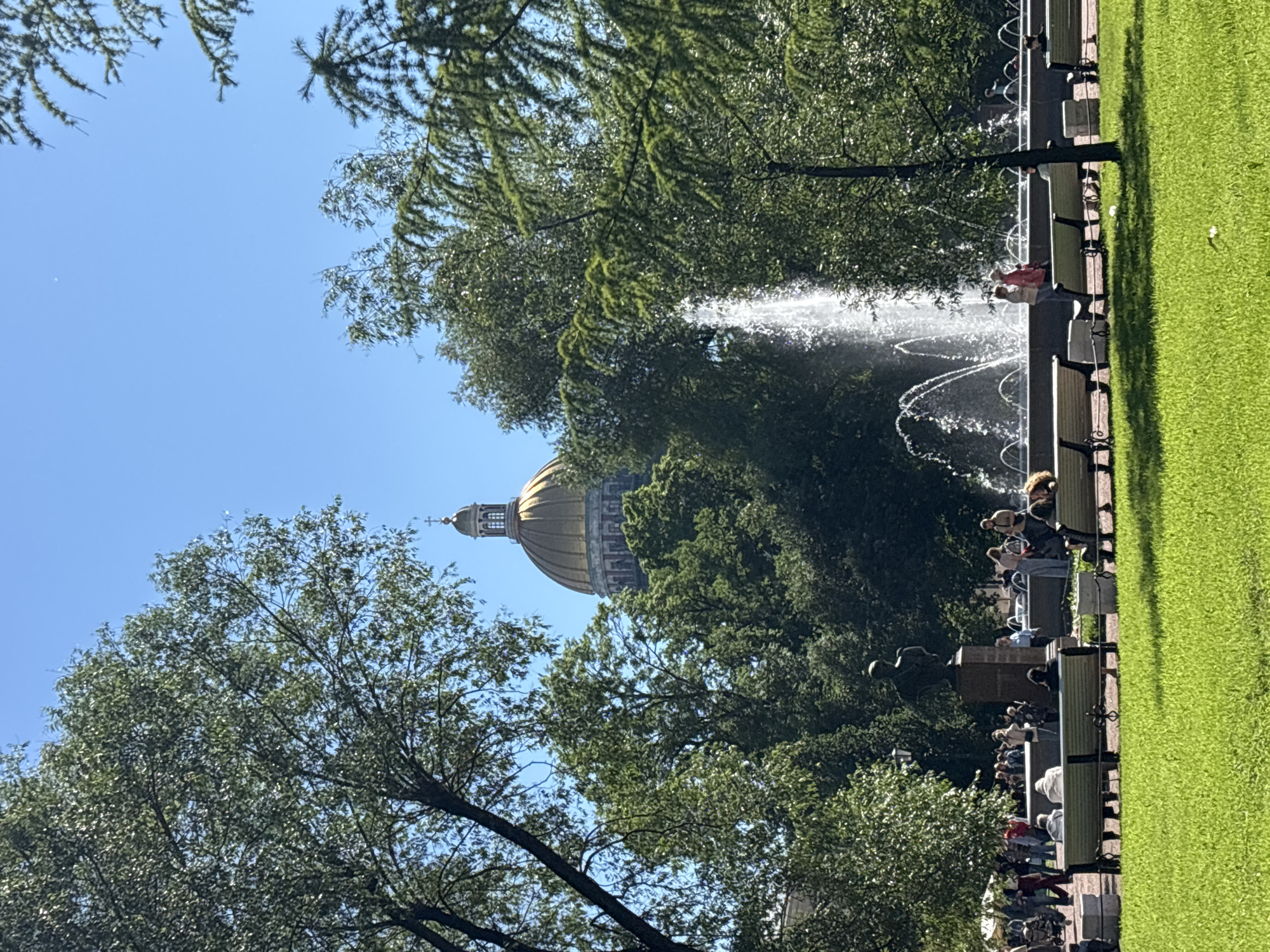 A clear, sunny day is depicted in a park with a fountain, benches, and trees. In the background, the golden dome of a building, possibly a cathedral, is visible. People are scattered throughout the park, some sitting on benches, others walking or standing near the fountain. The foreground features a lush green lawn.