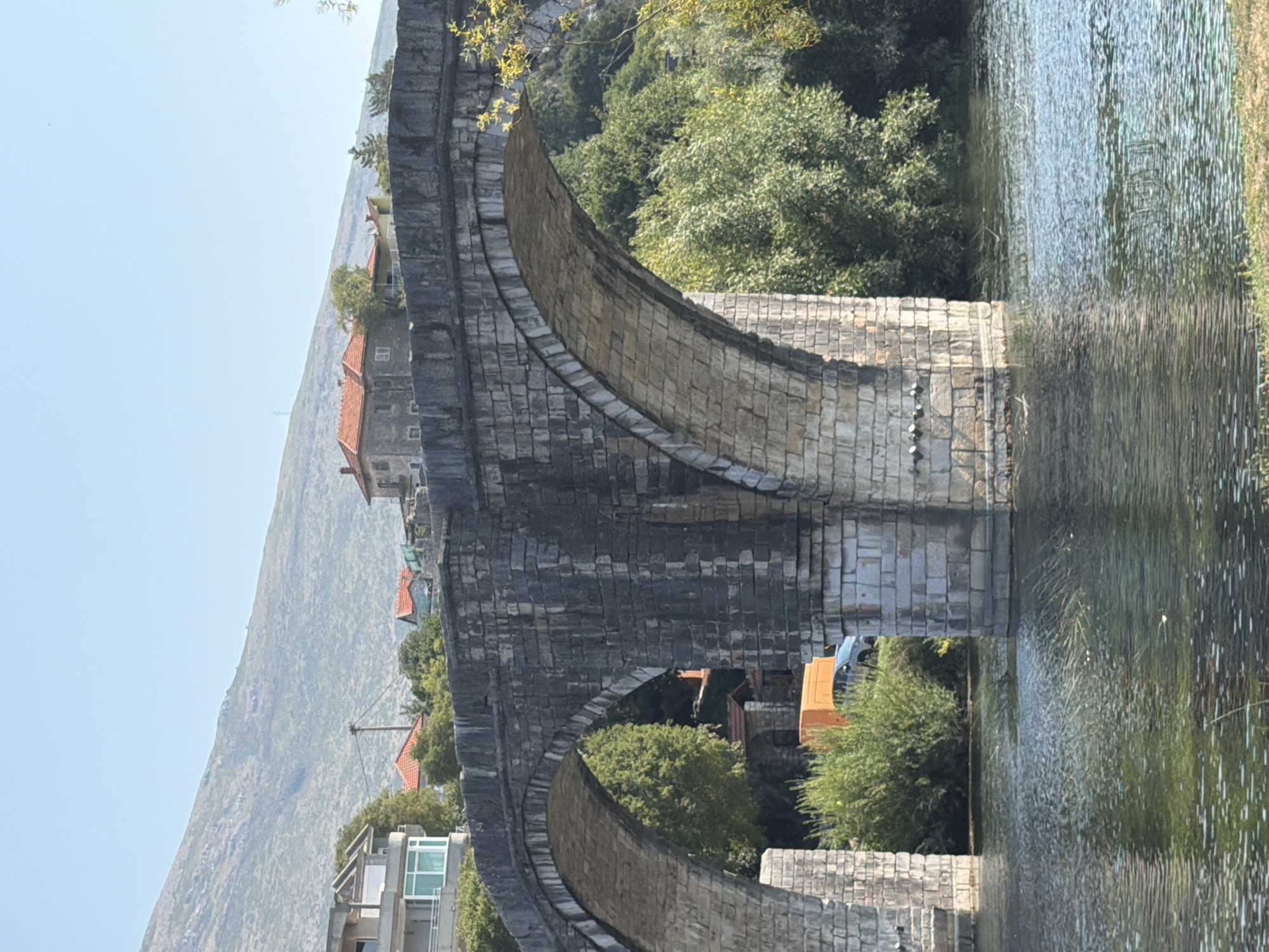 An ancient stone bridge with multiple arches spans a wide, calm river. The bridge is constructed from large, weathered stones, showing signs of age and erosion. Green foliage and trees grow densely along the riverbanks and behind the bridge. In the background, several houses with red tiled roofs are visible, and further back, a large, barren mountain slopes upwards under a clear blue sky. The scene appears to be the Arslanagić Bridge in Trebinje, Bosnia and Herzegovina.