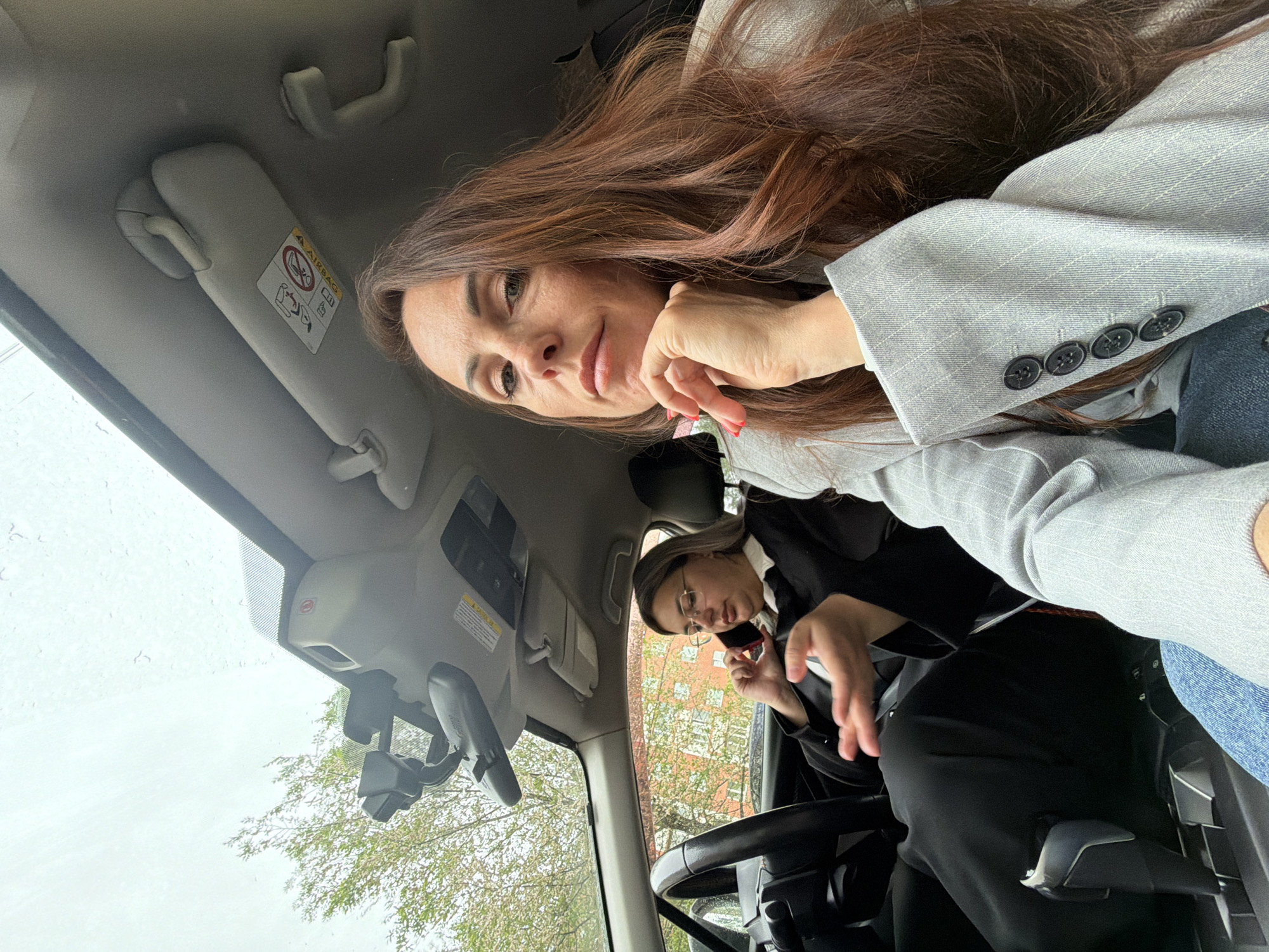 This image captures a moment inside a car, likely a selfie taken by the passenger from a low, angled perspective. Two women are present: one in the foreground and another in the background, appearing to be the driver.
The woman in the foreground, presumably the passenger, has long, flowing brown hair that cascades across her shoulders and chest. She is wearing a light grey blazer with subtle pinstripes over what looks like a white top, and blue jeans are visible on her lap. She is looking directly at the camera with a neutral to slightly smiling expression, her left hand gently resting near her chin, revealing red nail polish.
In the background, the driver is visible, wearing glasses, a black blazer, and a white shirt with black pants. She appears to be engrossed in a phone conversation, holding a red mobile phone to her ear.
The car's interior is visible, including the overhead console, a sun visor with a prominent "AIRBAG" warning label, and the steering wheel. Through the front windshield, an overcast sky is visible, along with some tree branches with green leaves and a distant building, suggesting an urban or suburban setting in Birobidzhan, Russia. The windshield also shows some moisture, possibly raindrops, indicating wet weather. The overall impression is of two people inside a vehicle, with one engaging in a photo and the other occupied with a call, during what appears to be daylight hours under a cloudy sky.