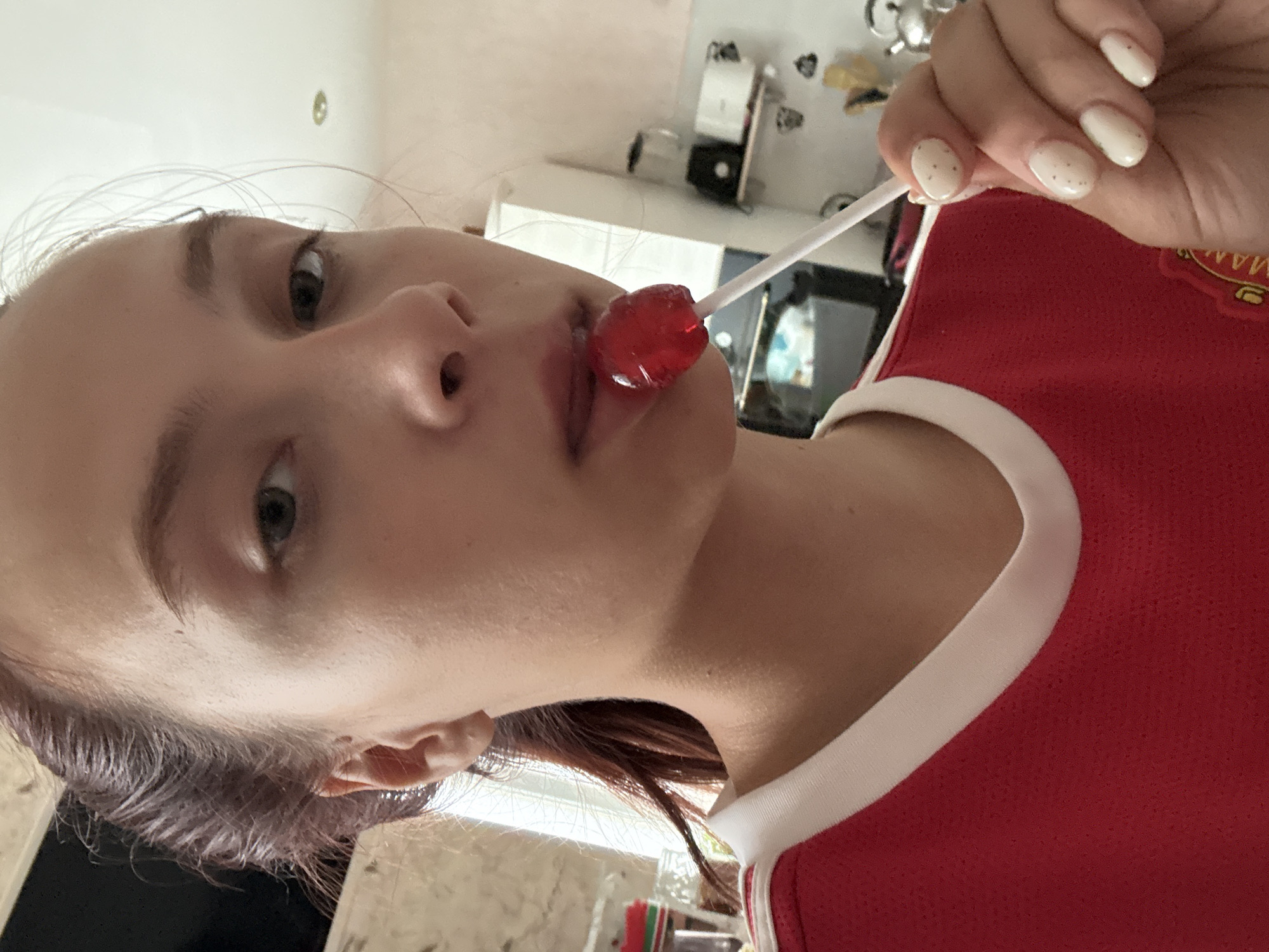 A young woman with brown hair pulled back is shown from the chest up, holding a red lollipop to her mouth. She is wearing a red shirt with a white collar. Her nails are painted white with small speckles. The background appears to be a kitchen with white cabinets and a microwave visible. The location is in the city of Perm, Russia.