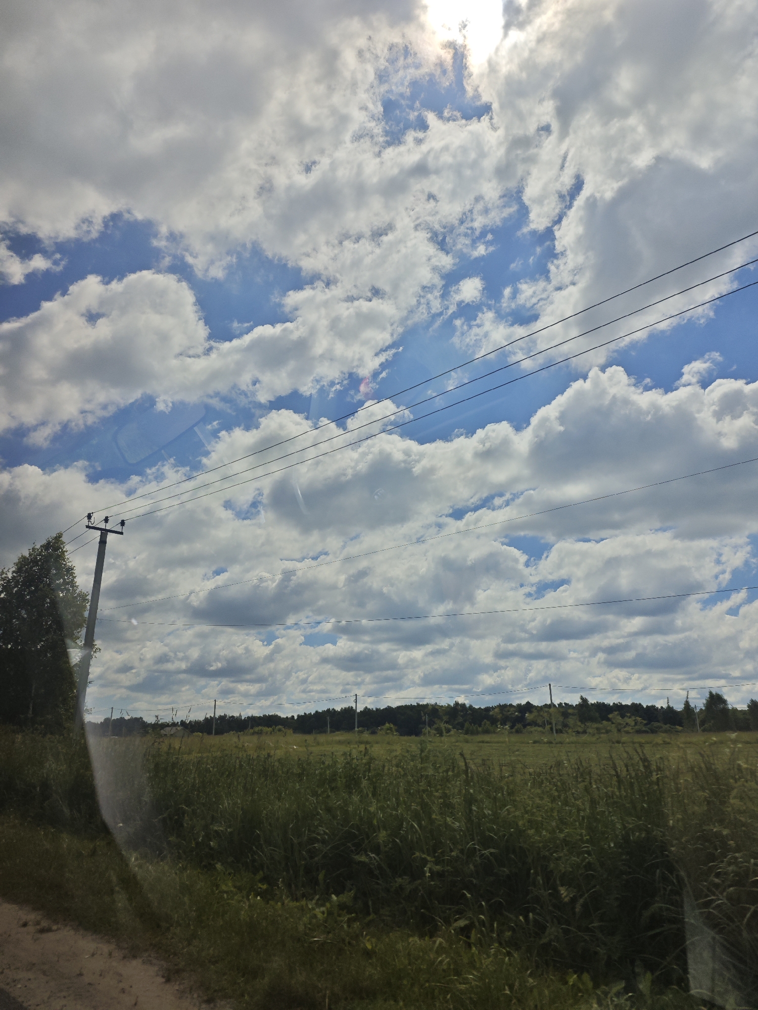The photograph captures a rural landscape under a bright, partially cloudy sky. The scene is dominated by a field of tall, green grass and a distant line of trees. In the foreground, a dirt road is visible on the left, partially obscured by vegetation. A telephone pole with wires stretches across the frame, adding a man-made element to the natural setting. The sky is filled with large, white cumulus clouds against a bright blue background, with the sun shining through, creating a bright, sun-drenched atmosphere. The image is likely taken from a vehicle, as indicated by a subtle reflection in the lower left quadrant. The location is identified as Klimovka, Russia.