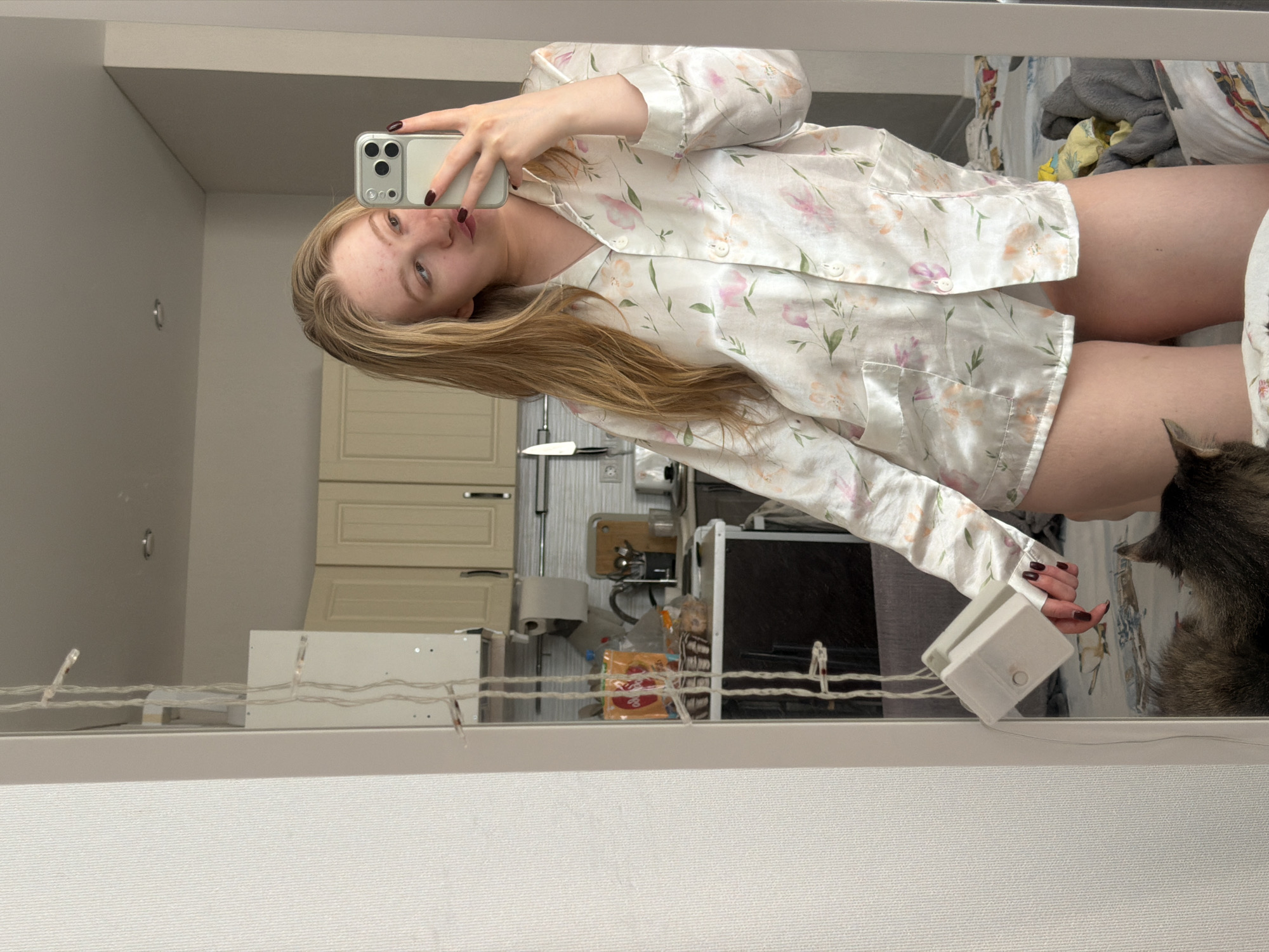 A woman is taking a selfie in what appears to be a mirror. She is wearing a white floral pajama top. Her long blonde hair is visible, and she is holding a cell phone in her left hand. The background shows a kitchen area with cabinets, various appliances, and some items on the counter. The reflection also includes a bed with various items, including what appears to be a cat. The location is Засечное, Russia.