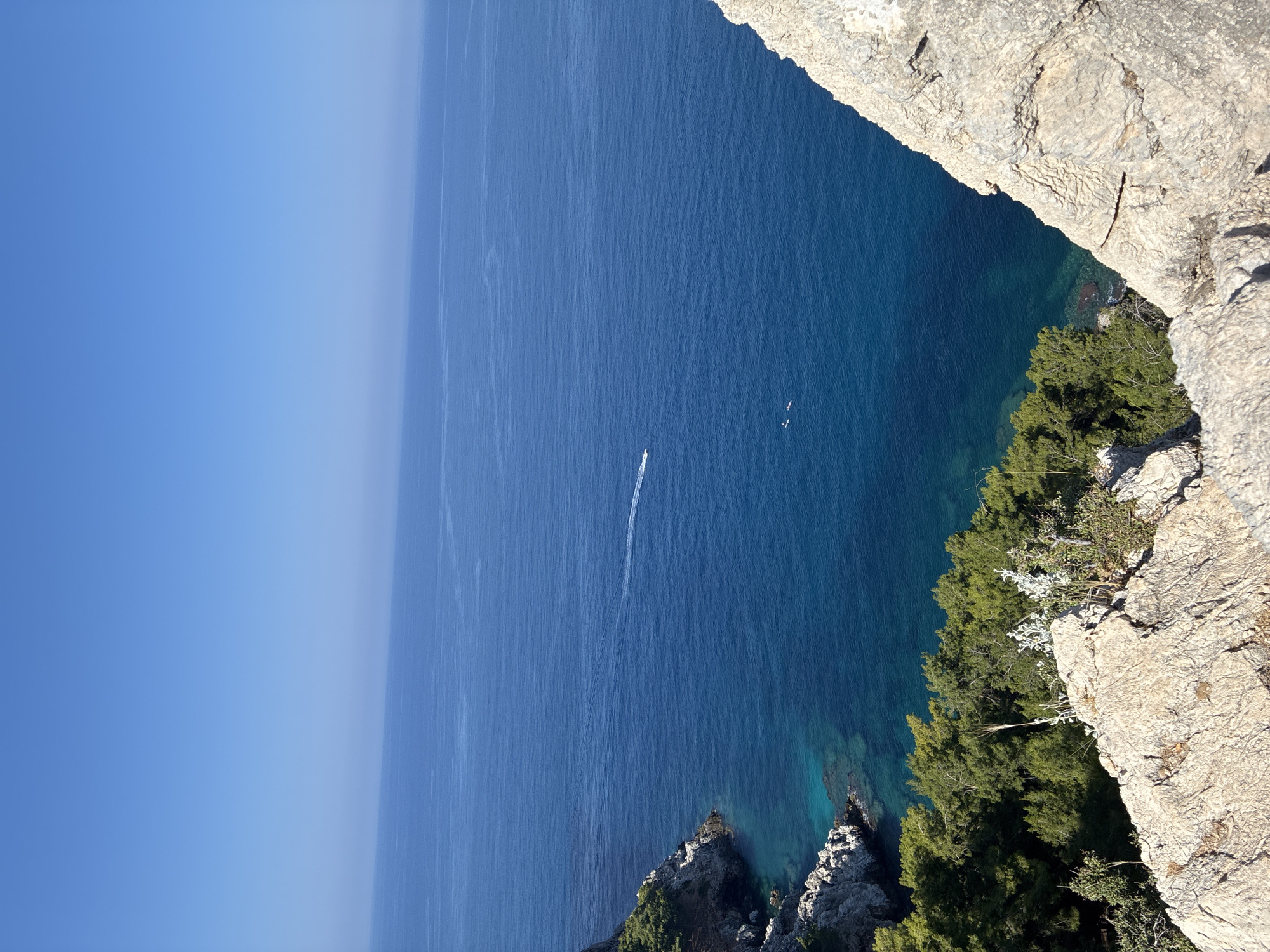The image depicts a scenic overlook of the Mediterranean Sea from a cliffside in Alanya, Turkey. The foreground is dominated by a rough, rocky cliff face with sparse vegetation. In the midground, a lush green treeline lines the edge of another cliff that drops down to the clear, turquoise water. The sea itself is a deep blue, stretching out to the horizon under a clear, pale blue sky. A white speedboat is visible in the distance, leaving a wake on the water. Two small figures, likely on paddleboards, are also present on the water. The overall impression is one of natural beauty and tranquility.