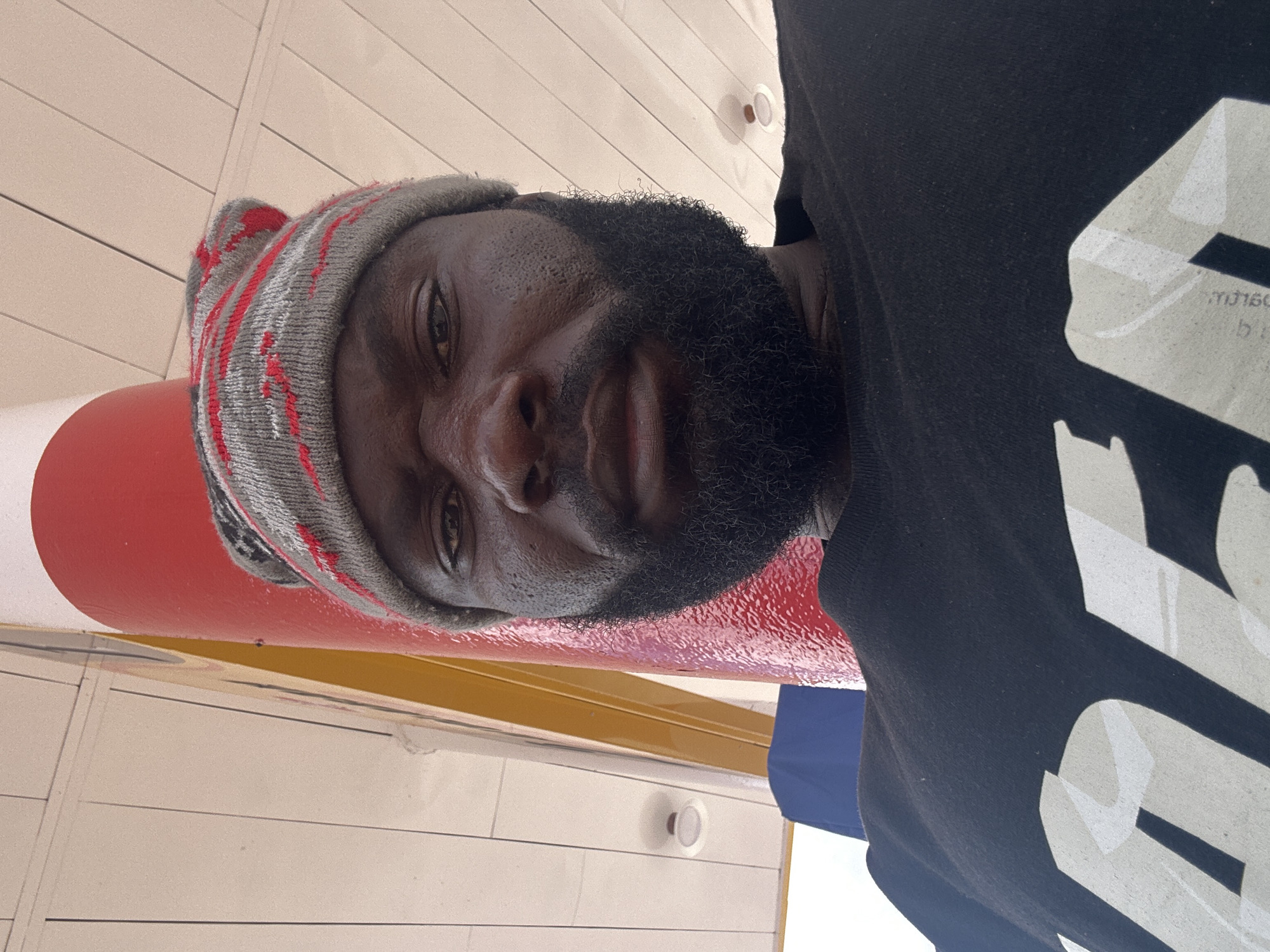 A man is captured in a selfie, facing the camera. He is wearing a gray and red striped knit beanie and a black t-shirt with large, light-colored block letters that appear to spell out "ART". His face is dark-skinned with a full black beard and mustache. He has sweat droplets visible on his forehead and cheeks. The background shows a bright red cylindrical pole and a ceiling with white panels and recessed lighting. The setting appears to be outdoors, possibly under an awning or at a gas station, with a hint of yellow structure to the left. The image was taken in Barakin Lerawi, Nigeria.