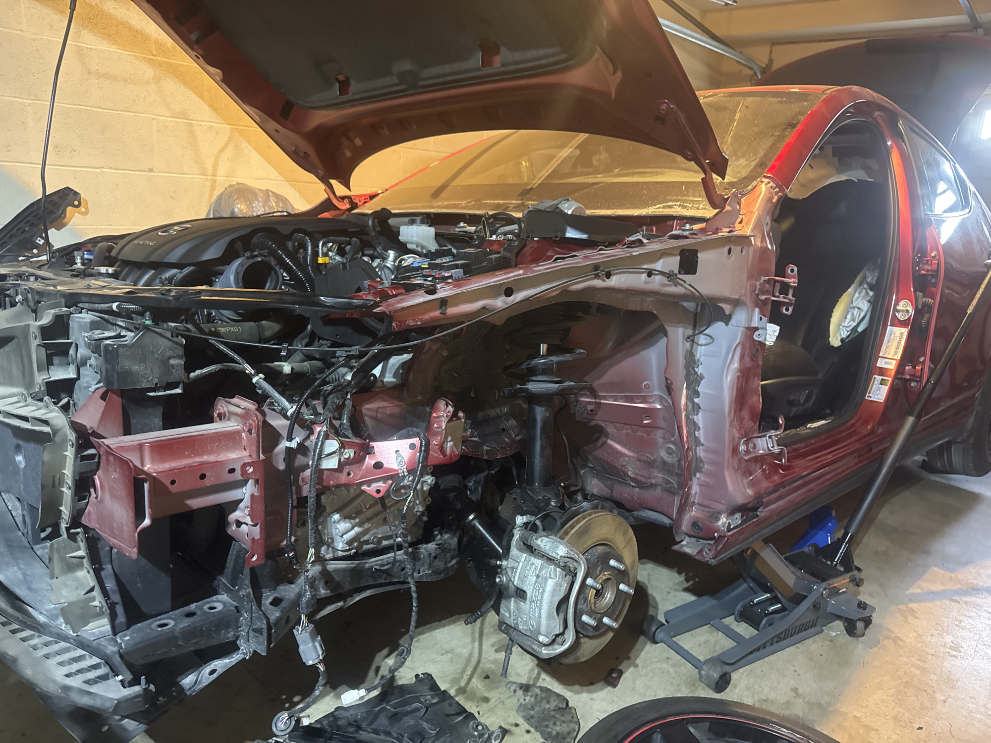 The image shows a dismantled red car in what appears to be a garage. The front of the car has been removed, exposing the engine and various mechanical components. The hood is open, revealing the engine bay. The car is supported by a hydraulic jack. The surrounding environment is a concrete floor with a textured wall in the background. The lighting suggests an indoor setting, possibly a workshop or garage.