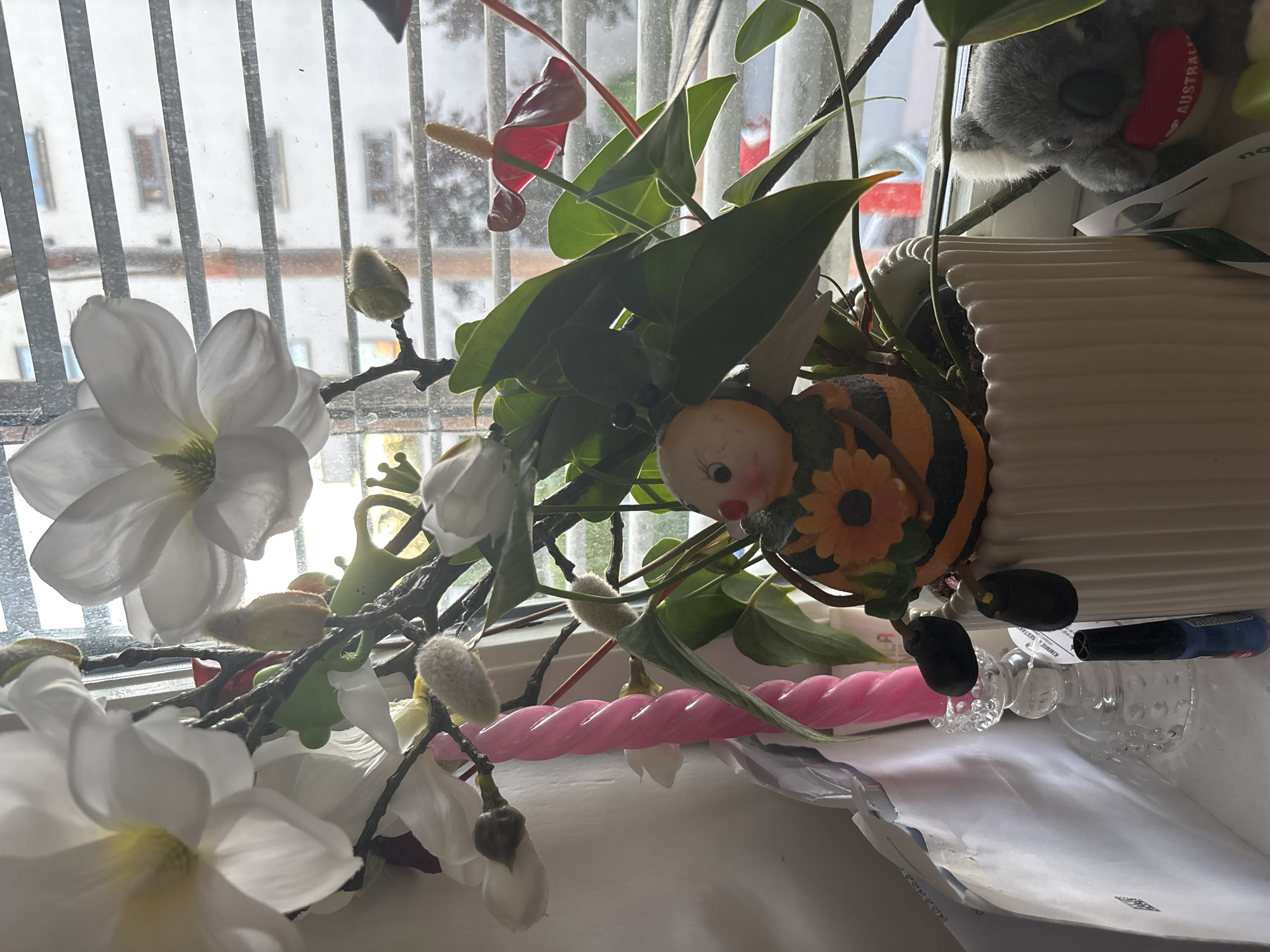 An indoor scene is depicted with a variety of decorative objects and plants placed near a window with blinds. In the foreground, a white fluted planter holds a decorative bee figurine with black and orange stripes and a yellow sunflower on its body. The bee has black legs and a round face with eyes and red cheeks. Next to the planter, a pink twisted candle stands in a clear glass candlestick holder. To the left of the candlestick, a stack of white envelopes is visible. To the right of the planter, a small plush koala wearing a red heart with the word "AUSTRALIA" is positioned. Behind these items, artificial white magnolia flowers and green leaves are arranged. A red anthurium flower with a yellow spadix is also present. The background shows a window with horizontal blinds, through which a blurry view of buildings and trees can be seen. The overall lighting suggests an overcast day. The scene appears to be a domestic setting, likely a windowsill or shelf.