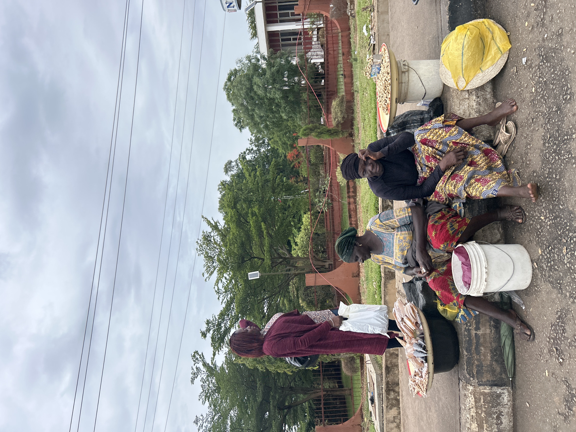 Three women sit on a sidewalk selling items. One woman in a maroon dress stands looking at the items being sold. The women who are selling are sitting on the ground next to various containers including buckets, bowls, and a yellow straw hat. The items being sold include what appear to be nuts and fish in plastic bags. In the background is a red metal fence and trees. Power lines are visible in the sky. Based on the style of clothing and the background, this appears to be in Jos, Nigeria.