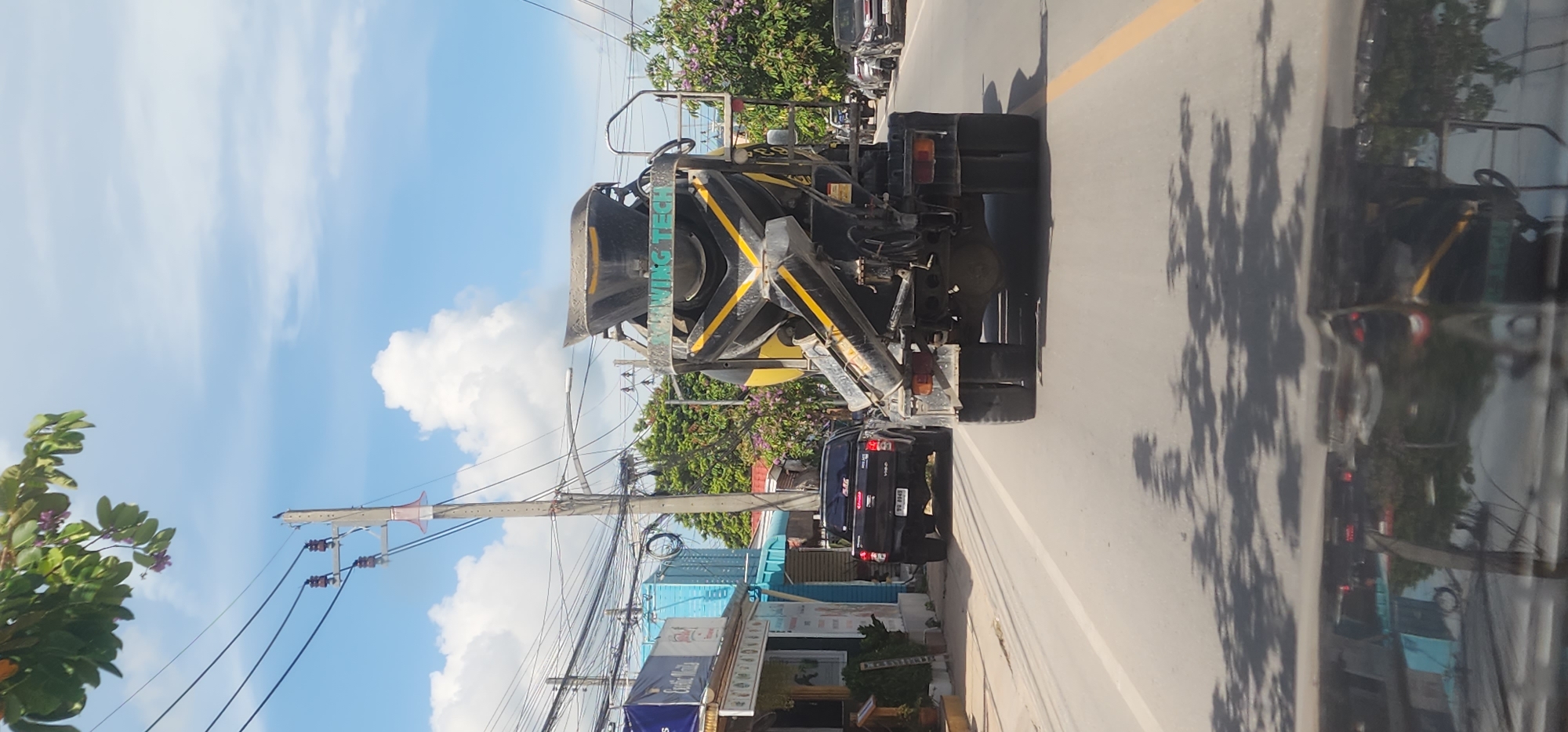 A cement truck is driving on a road in Baan Plai Laem, Thailand. The truck is black and yellow and has a large mixing drum on the back. A black pickup truck is in front of the cement truck. The road is lined with buildings and trees. The sky is blue with some clouds. The image appears to be taken from another vehicle, as evidenced by the reflection on the hood.