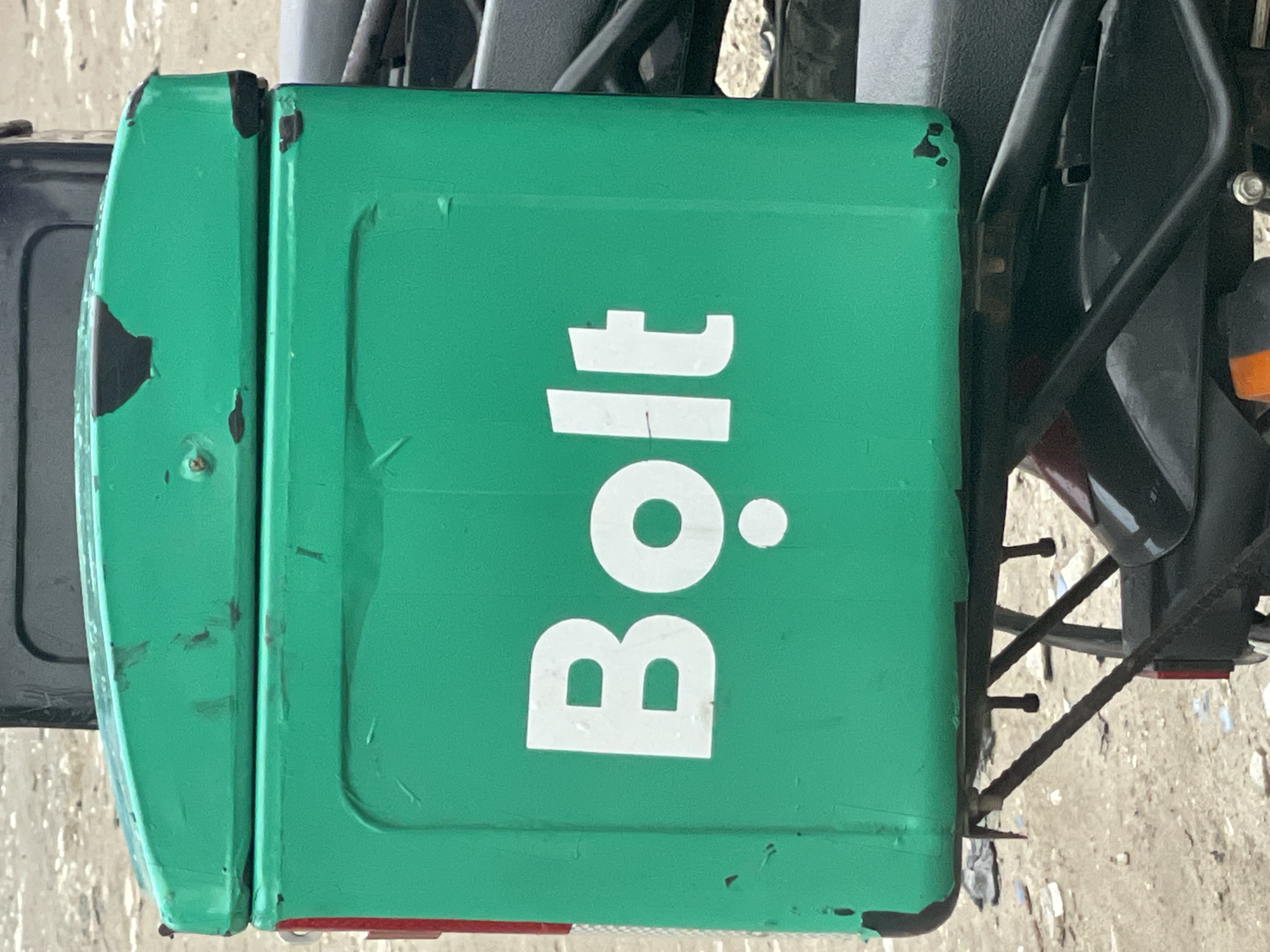 A close-up, eye-level, medium shot captures a green delivery box attached to the back of a motorcycle. The box has the white logo "Bolt" printed on it, with a small white circle below the 'o'. The box shows signs of wear, with scuffs and some chipped paint, particularly on the top edge. The background is out of focus and appears to be a sandy or dusty surface, suggesting an outdoor urban environment. Part of the motorcycle seat and frame are visible below the box. The image was taken in Ilado, Nigeria.