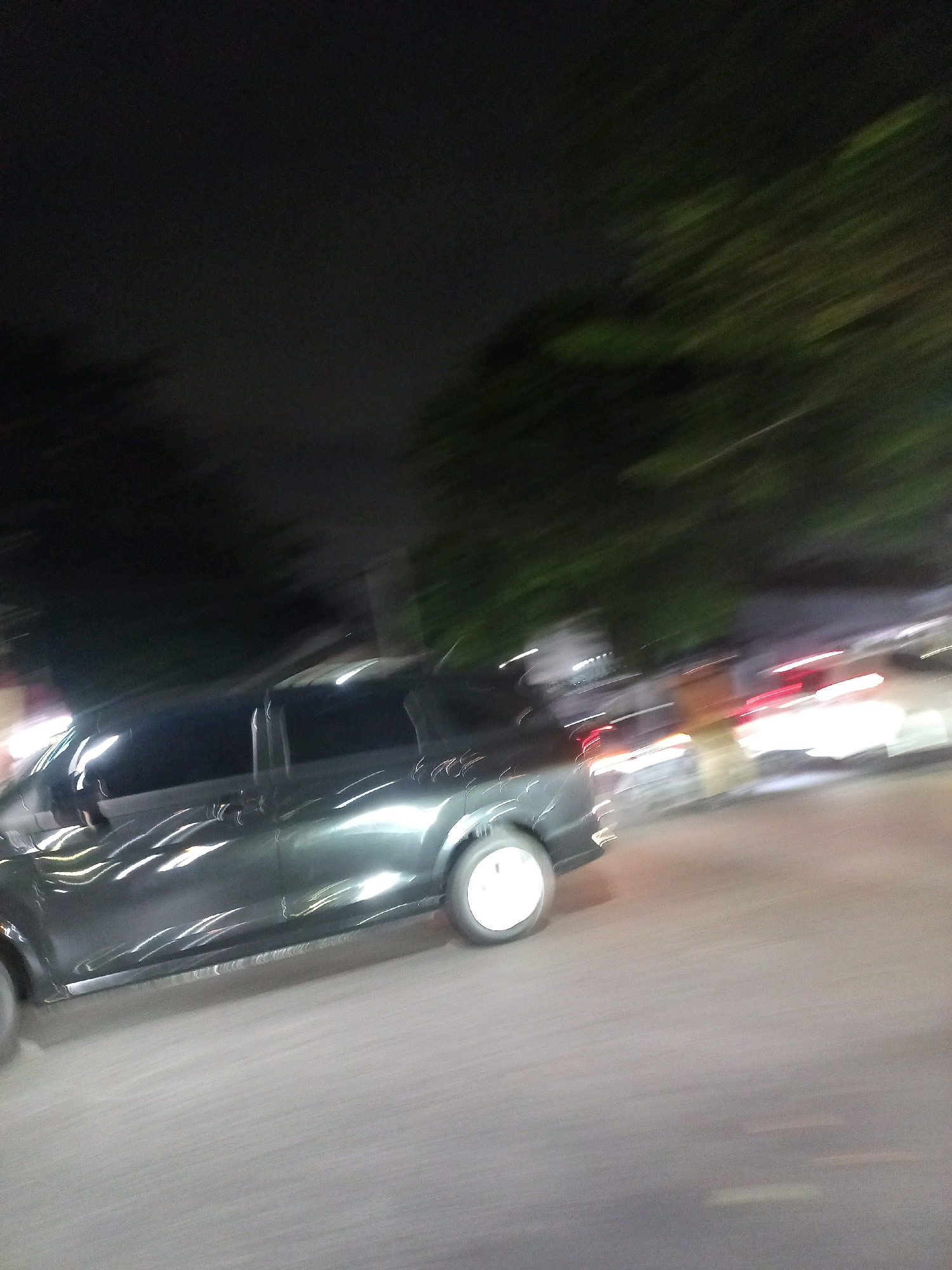 The image captures a motion-blurred scene at night, likely on a street in Depok, Indonesia. A dark-colored car, possibly a hatchback or small SUV, is the primary subject, moving from left to right. The car's surface reflects streaks of light, indicating streetlights or other vehicles. Its rear wheel is visible, and it appears to be a silver alloy wheel. The car's windows are tinted. In the background, blurred trees with some green foliage are visible against a dark sky. Further to the right, the taillights of another vehicle are visible as red streaks, suggesting it is also in motion or stationary. The road surface is asphalt, with some faint lighter-colored markings. The overall impression is a snapshot of urban traffic at night, with the motion blur conveying a sense of speed or movement.