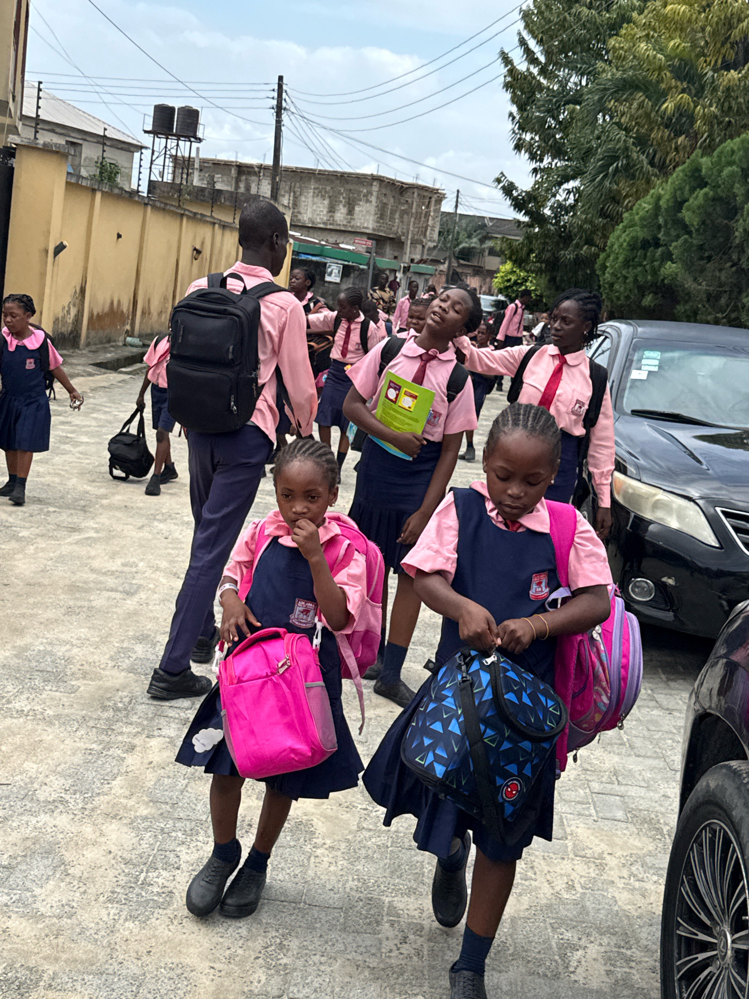 In the city of Ado, Nigeria, a group of schoolchildren in navy blue uniforms with pink shirts and ties are gathered on a paved street. The children are carrying backpacks and bags, some of which are bright pink. One young girl in the foreground is looking down at her bag, while another, slightly behind her, is walking and holding a pink backpack. A teenage boy in the background wears a pink shirt and navy pants, carrying a large black backpack. A black car is parked on the right side of the image. The background shows a yellow wall, buildings, and overhead power lines. The sky is overcast.