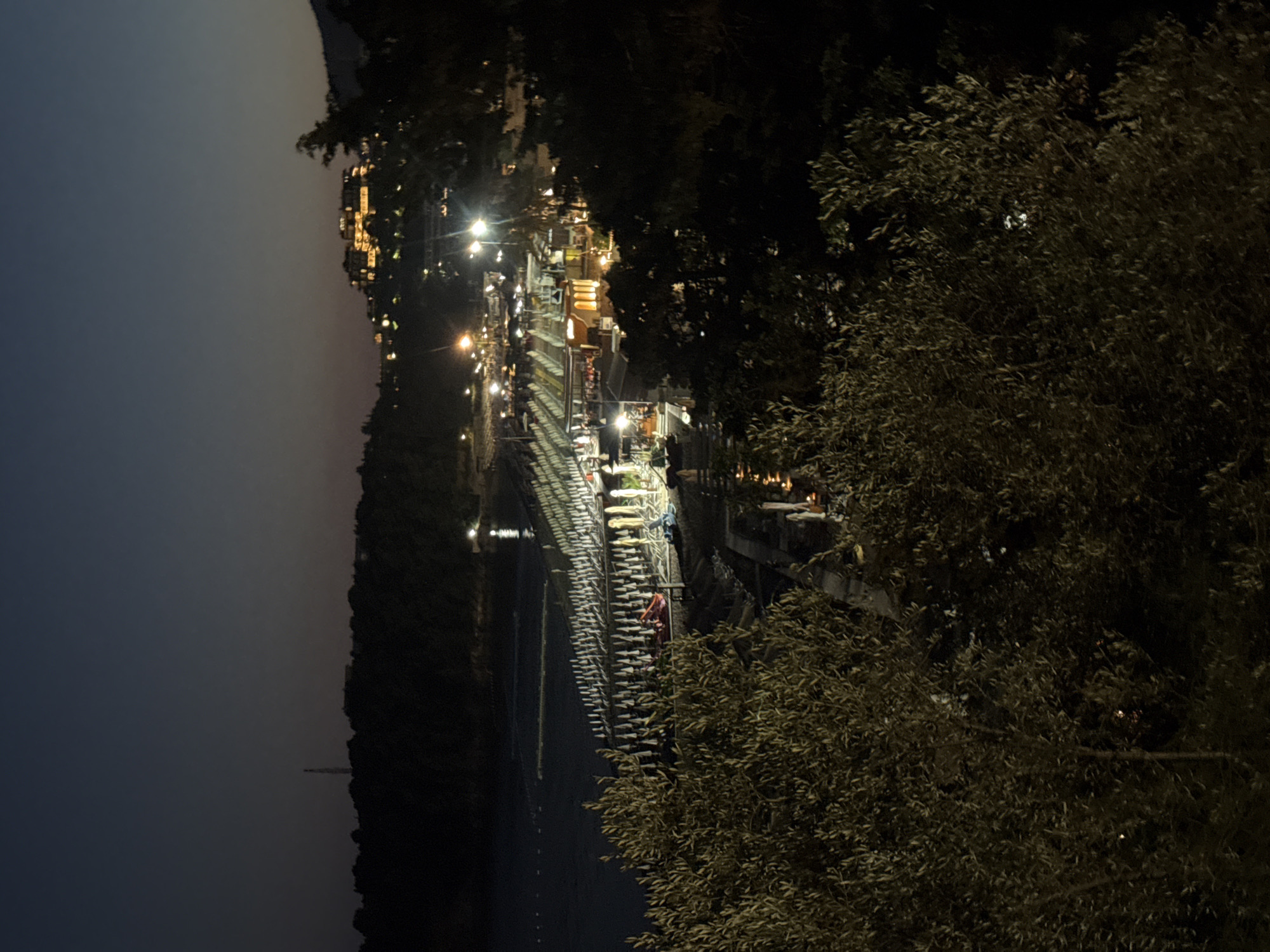 The image depicts a nighttime view of the shoreline and a beach in Boreti, Montenegro. The scene is illuminated by artificial lights from a resort or hotel complex visible on a hill in the background. In the foreground, lush green trees with olive-like leaves dominate the frame.
The beach itself is lined with rows of white beach umbrellas and chairs, suggesting it is a developed tourist area. A body of water, likely the Adriatic Sea, is visible to the left of the beach, with calm conditions and some distant lights reflecting on its surface. Several structures, possibly beach bars or restaurants, are also lit up along the beachfront, with patrons visible in some areas. The overall atmosphere is serene and illuminated, indicative of a resort town at night.