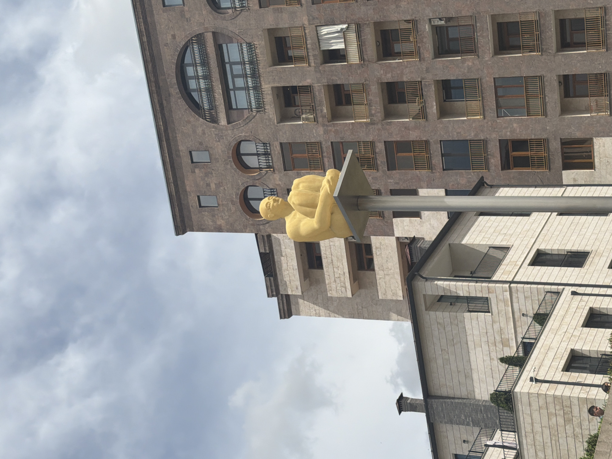 The image depicts a yellow sculpture of a man resting on a square platform atop a pole in Yerevan, Armenia. The sculpture is positioned in front of two multi-story buildings with a cloudy sky in the background. The building behind the statue has a stone facade with round windows and balconies, while the building to the right has a lighter-colored facade with square windows and balconies.
