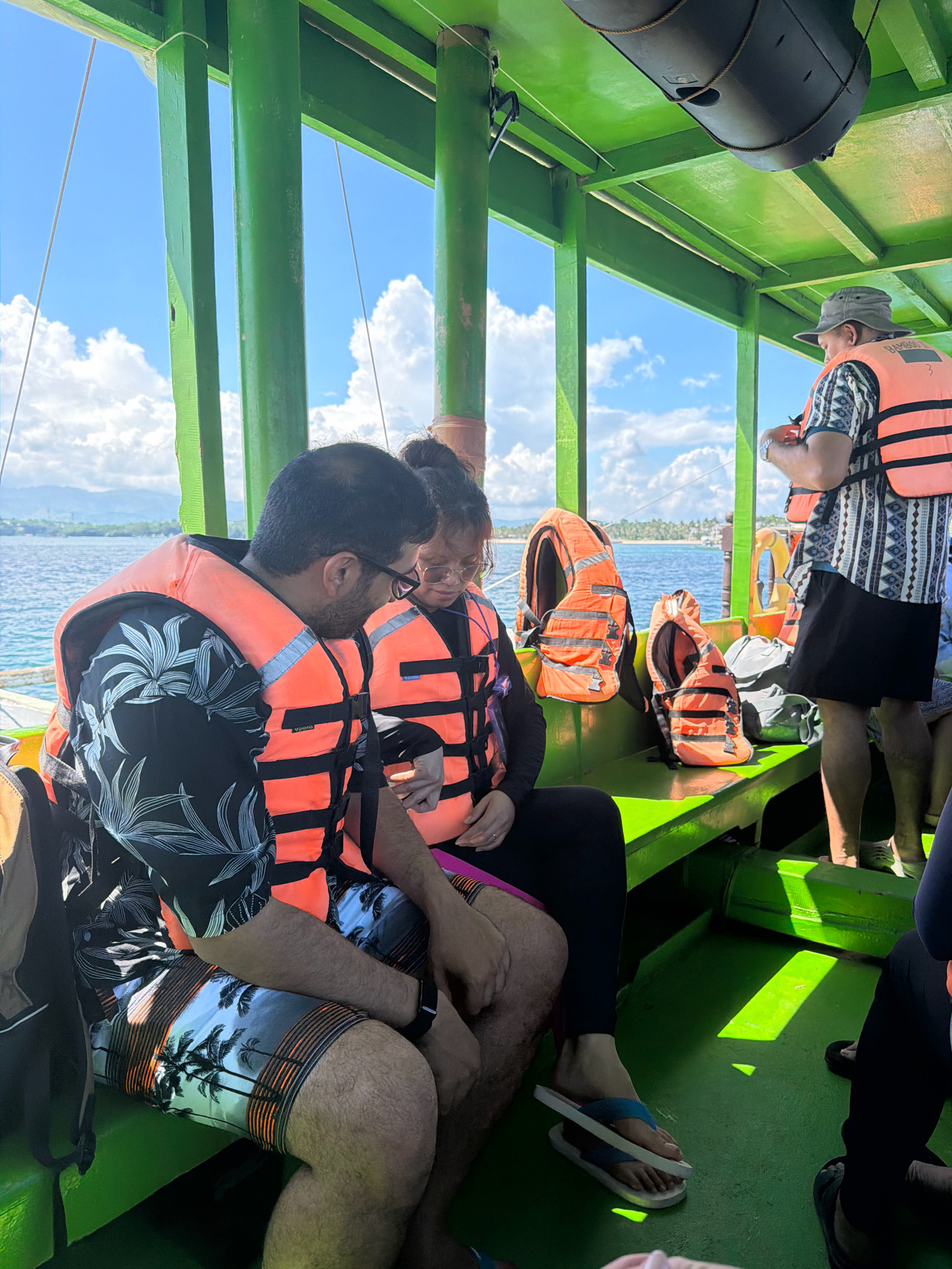 The image shows a group of people wearing orange life vests inside a green boat, likely traveling in the waters near Malay, Philippines. In the foreground, a man and a woman are seated, both wearing life vests and looking down. The man is wearing a black shirt with a floral pattern and patterned shorts. The woman is wearing a dark long-sleeved shirt and black leggings. Behind them, another person in a patterned shirt and dark shorts stands, holding onto something. Several orange life vests are stacked on a green bench in the middle of the boat. The boat's interior is painted green, with vertical support beams visible. Through the open sides of the boat, a bright blue sky with scattered white clouds and a distant coastline with vegetation can be seen. The water is calm and appears to be a tropical sea. The overall impression is of a scenic boat ride in a tropical destination.