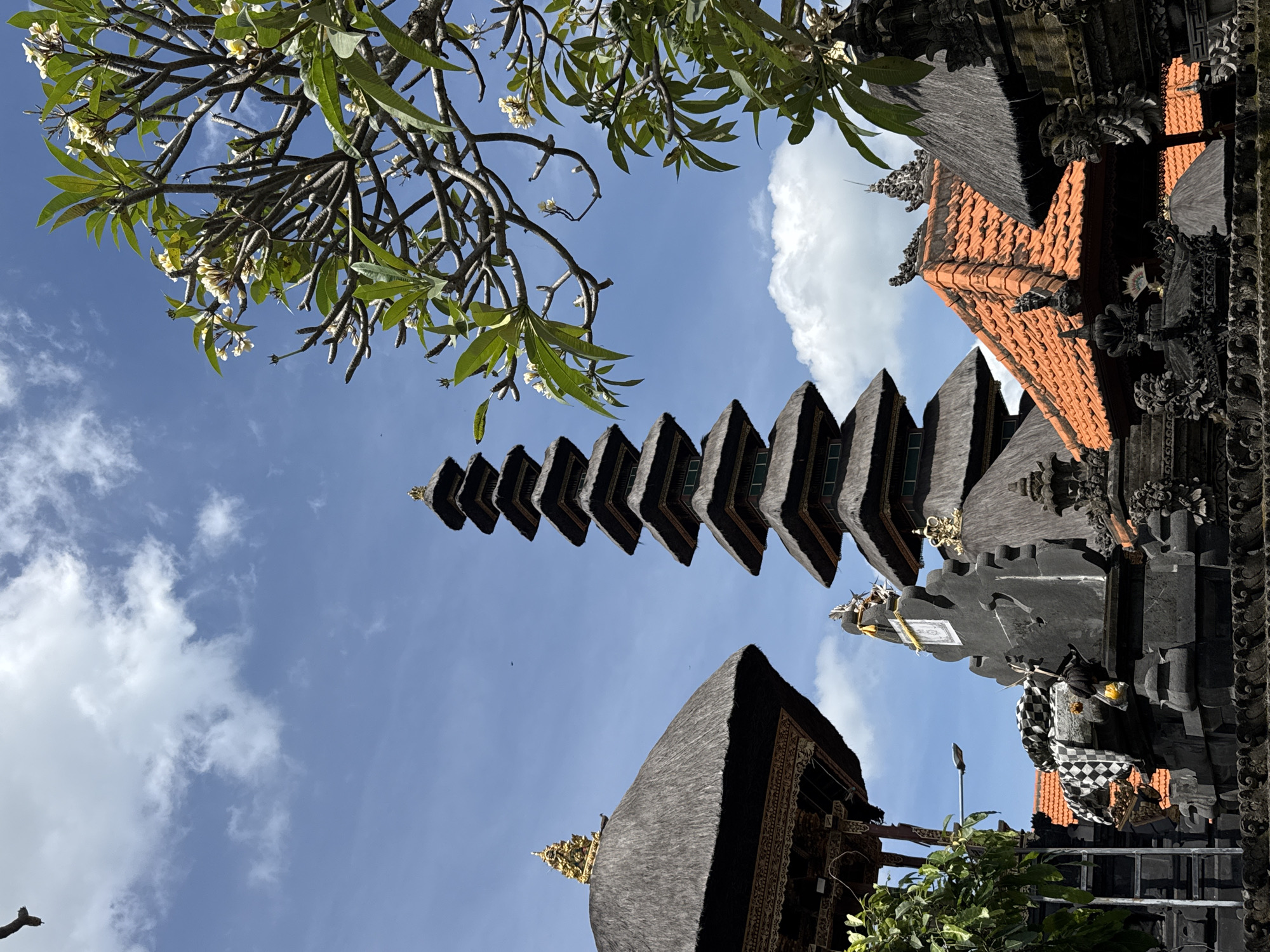 The image captures a low-angle view of a Hindu temple complex in Mengwi, Indonesia, seen against a blue sky with scattered clouds. The architecture features tiered pagoda-like structures with dark, layered roofs, as well as other buildings with thatched and tiled roofs. Elaborate carvings and decorations adorn the structures. Frangipani trees with green leaves and small white flowers frame the upper left portion of the image.