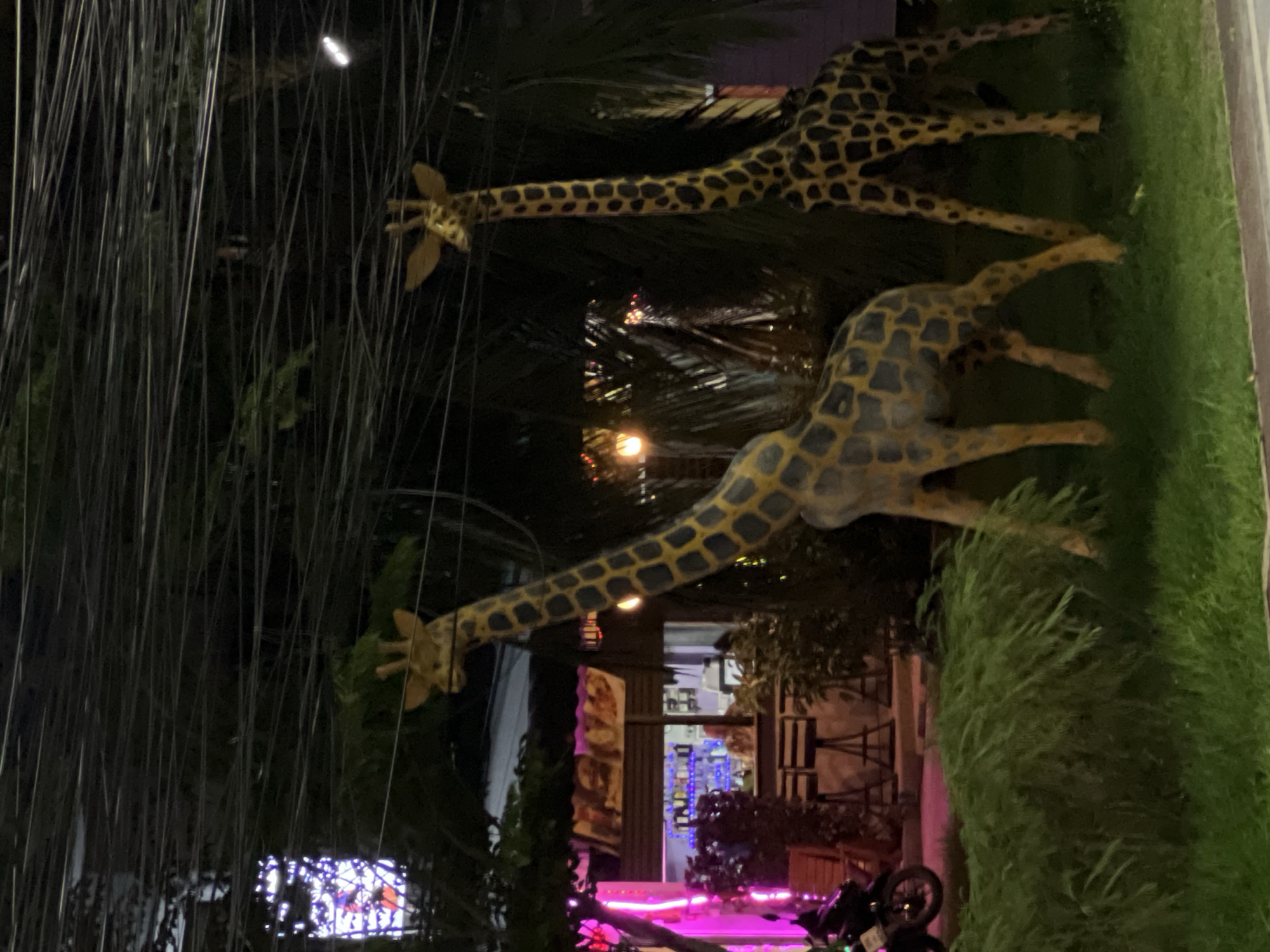 Two giraffe statues stand next to a shop in an unknown city. The statues are made of a tan material with brown spots. The shop is lit with bright neon lights and has a motorcycle parked in front. The background is obscured by trees and wires.